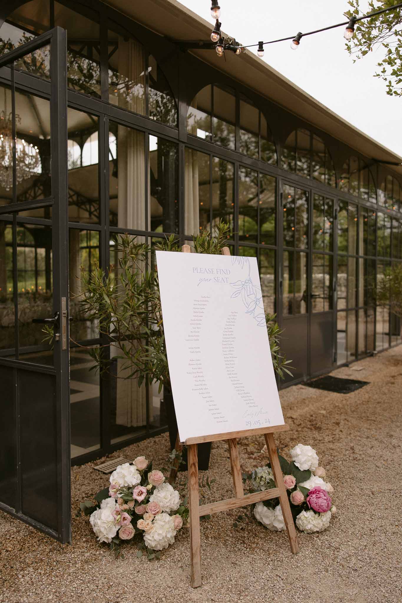 Seating chart sign on wooden easel with hydrangea and rose arrangements in front of glass-walled venue