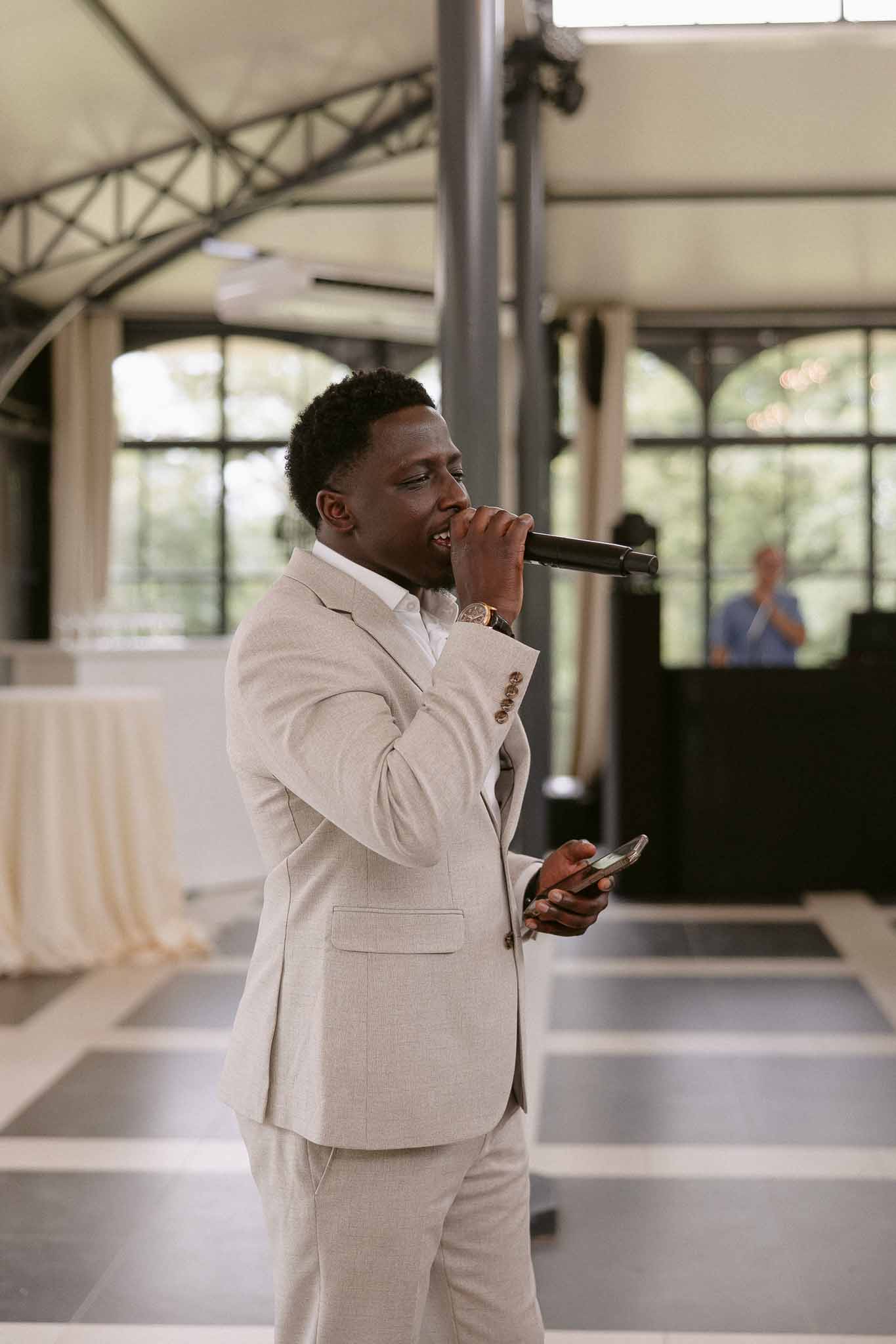 MC in beige suit speaking into microphone on checkered dance floor under white tensile roof with arched windows