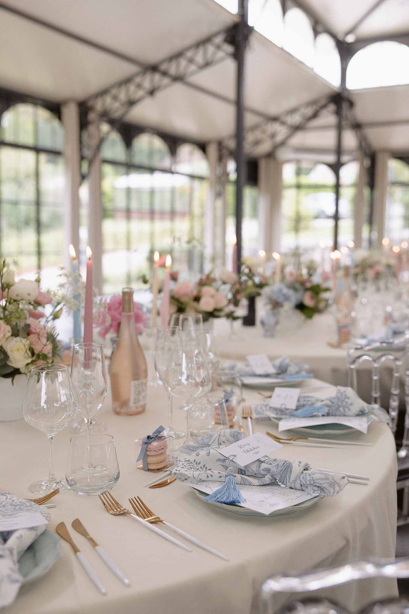 Reception table with toile napkins, gold cutlery, pink macaron favors, and blush rose centerpieces in iron orangery