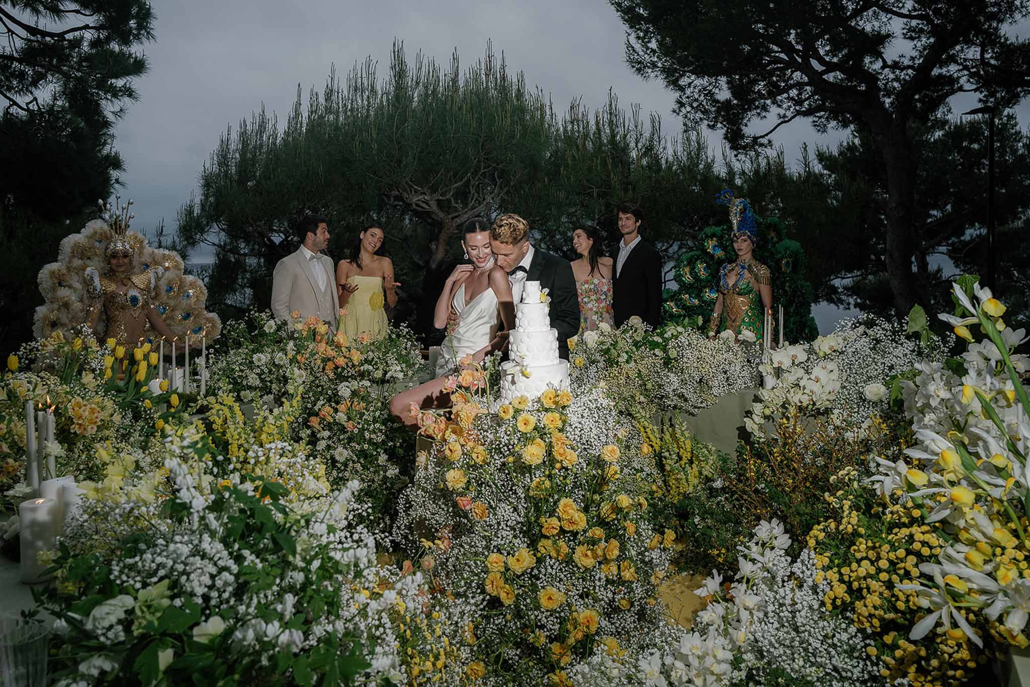 Couple cutting five-tier white cake amid yellow tulip and rose installation with costumed performers at dusk