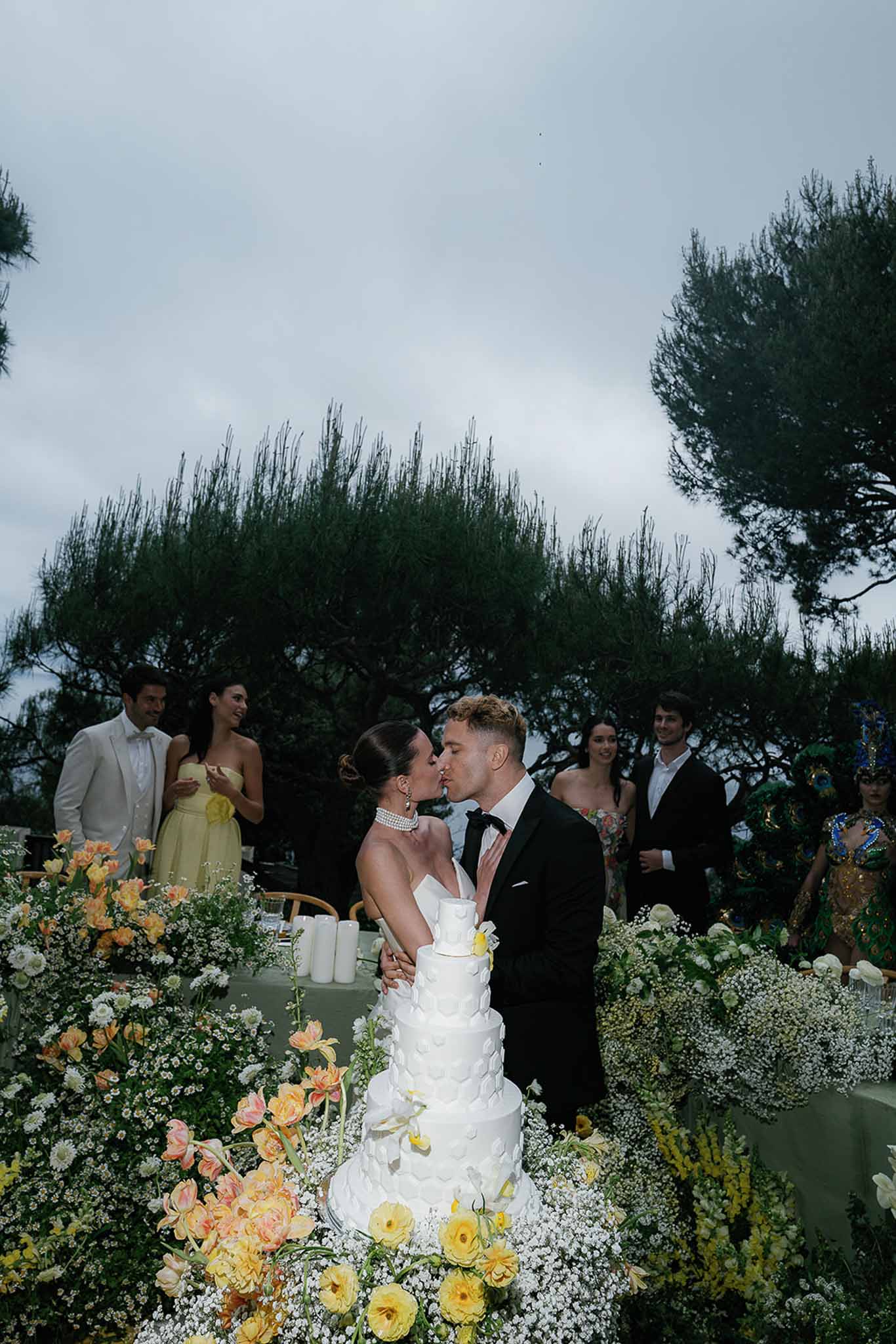 Couple kissing beside five-tier white cake surrounded by peach tulips, yellow roses, and mimosa installation