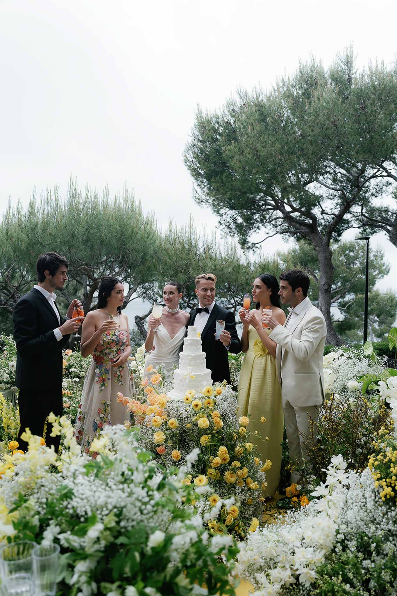 Couple and guests toast beside four-tier cake with yellow ranunculus and white gypsophila ground display