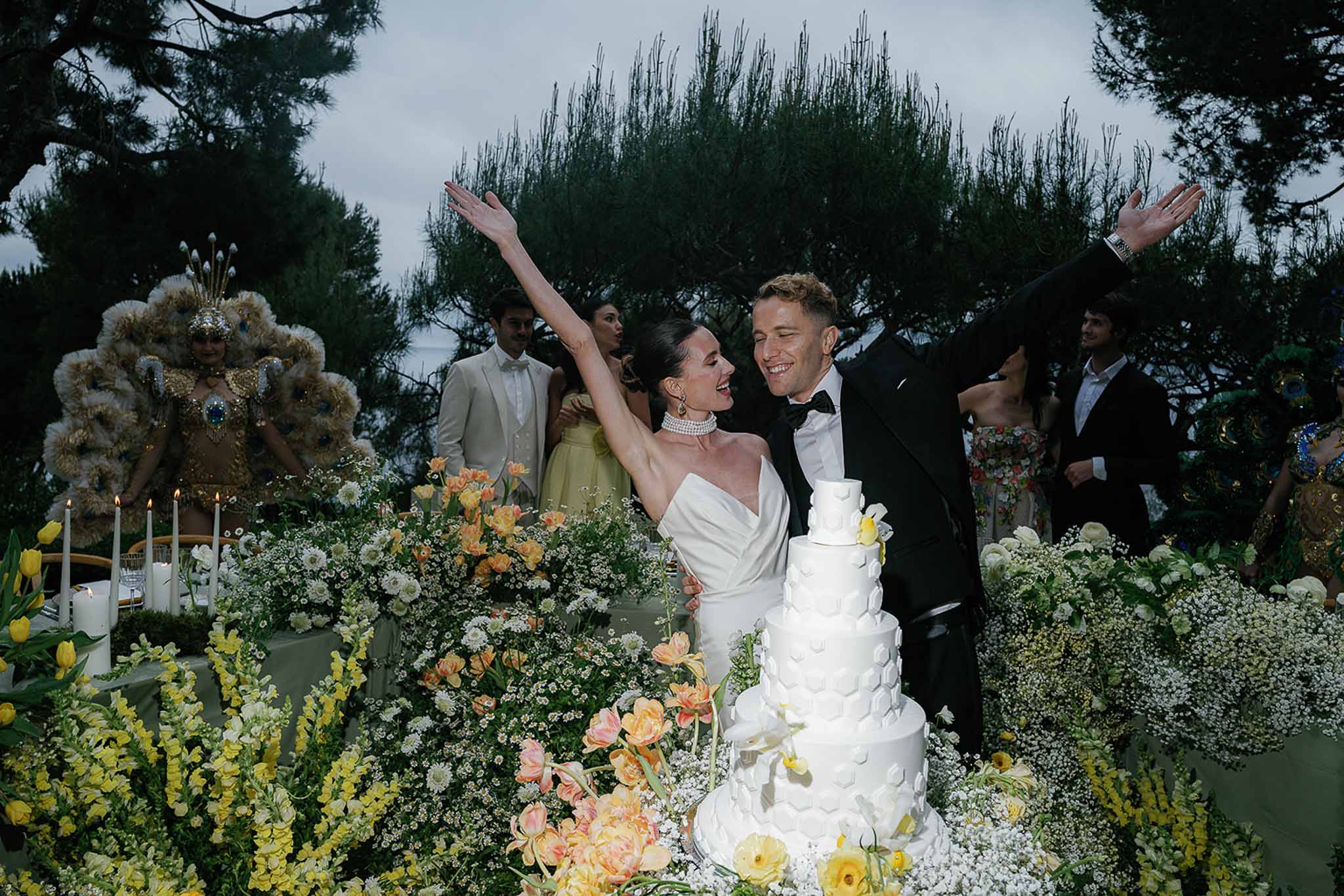 Couple celebrating at five-tier petal cake with peach tulips and costumed performers on evening terrace
