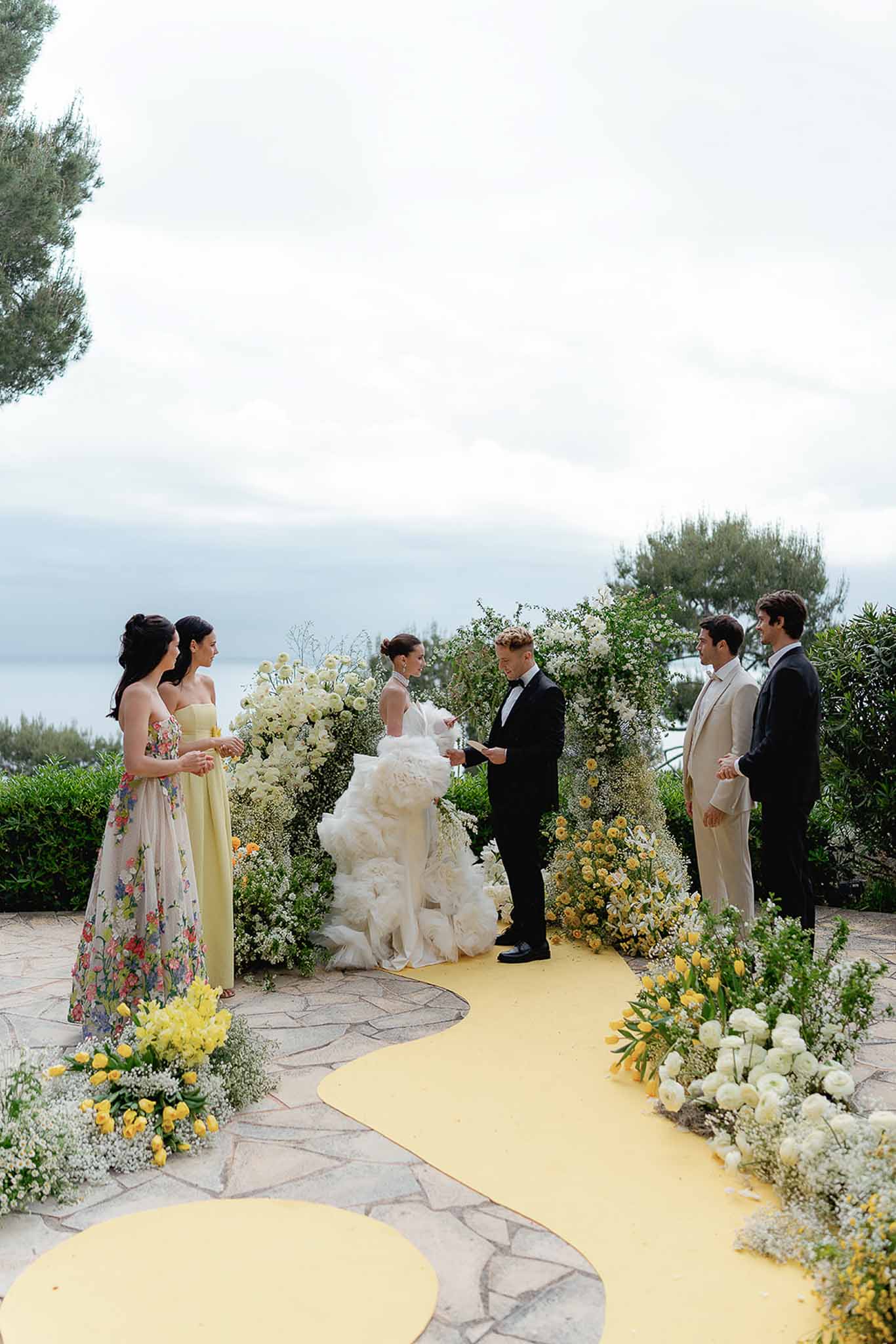 Seaside ceremony with yellow aisle runner, greenery arch with yellow tulips, and bridesmaids in yellow and floral gowns