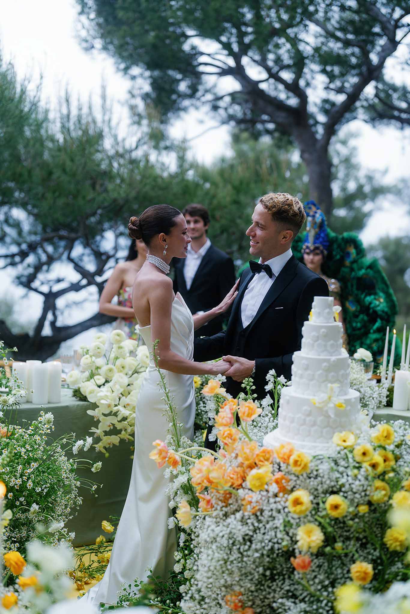 Bride and groom holding hands beside five-tier cake and yellow and orange floral installation at outdoor reception