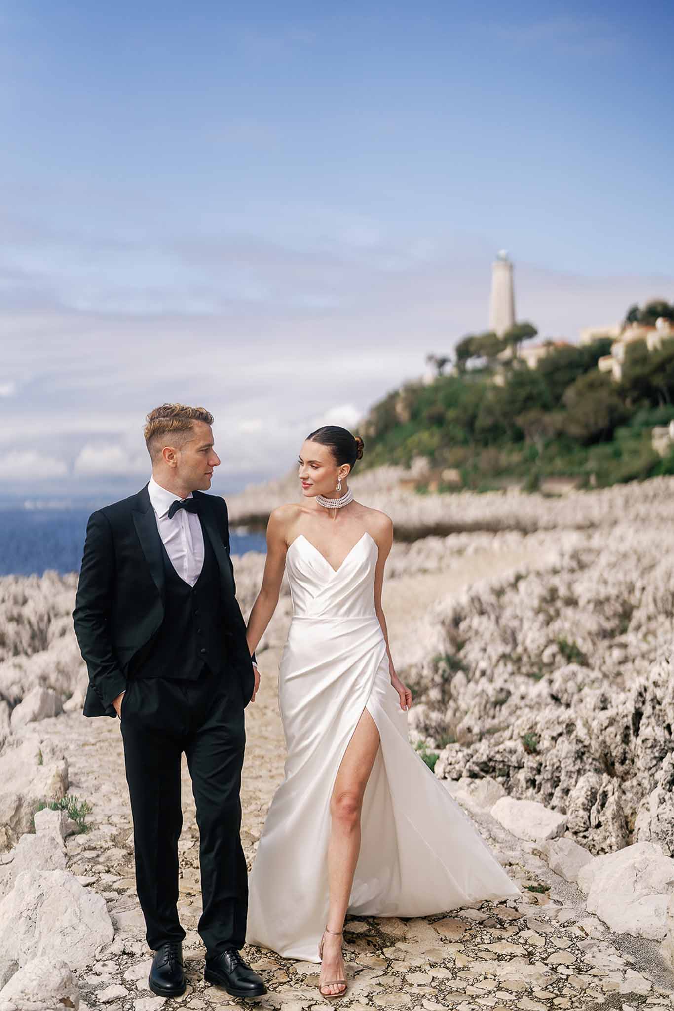 Couple walking on rocky coastal clifftop bride in strapless satin wrap gown groom in three-piece tuxedo lighthouse behind