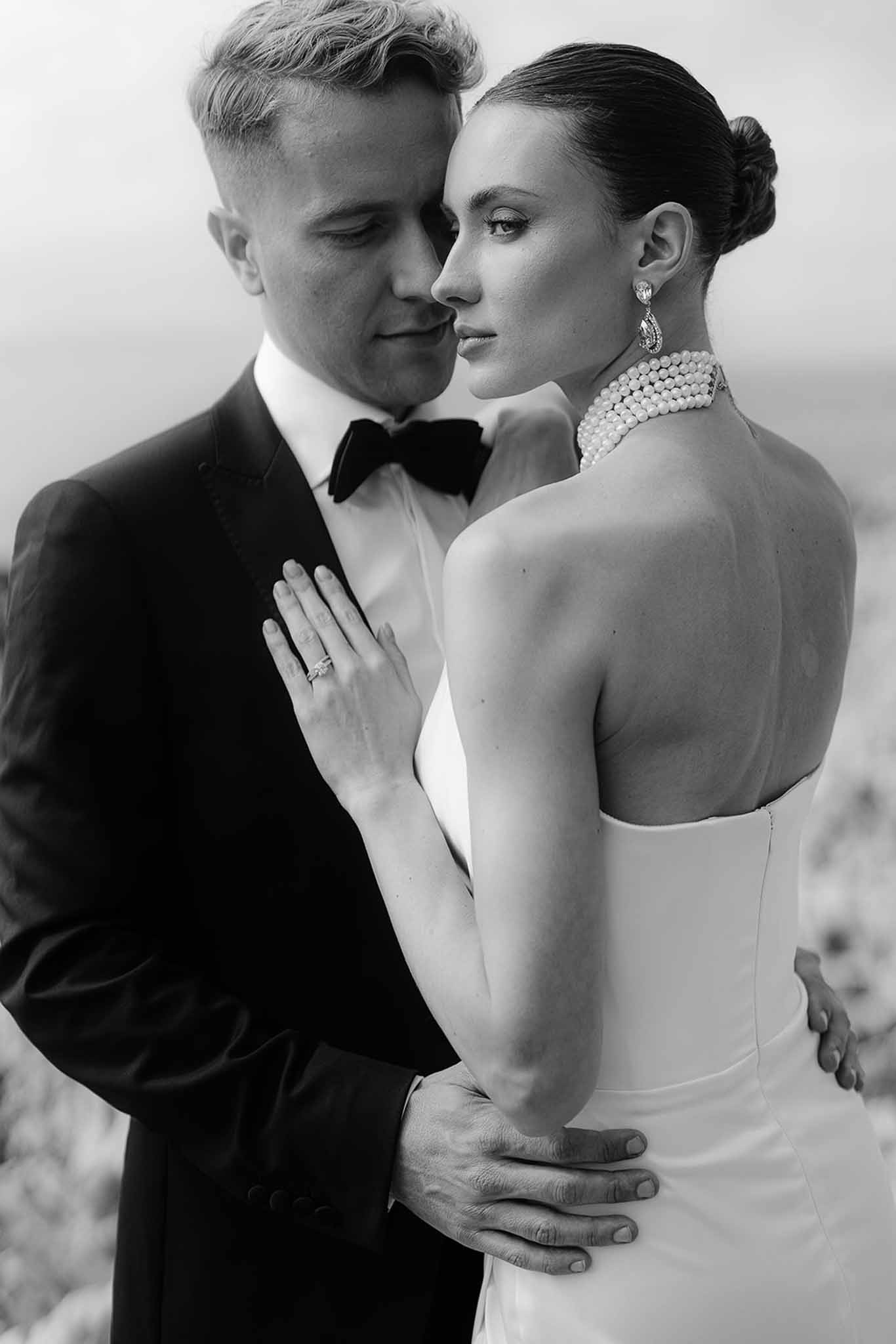 Close-up of bride's open-back gown and pearl choker with groom's arm around waist in editorial B&W