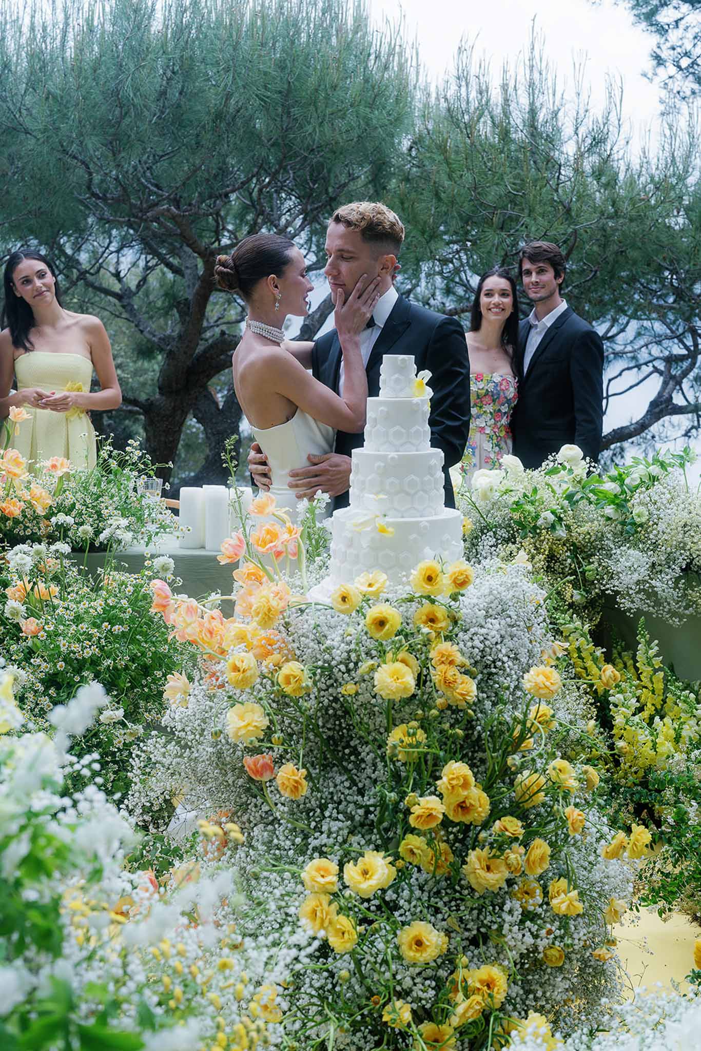Bride and groom share a moment during cake cutting beside a five-tier white cake and yellow and peach floral arrangement