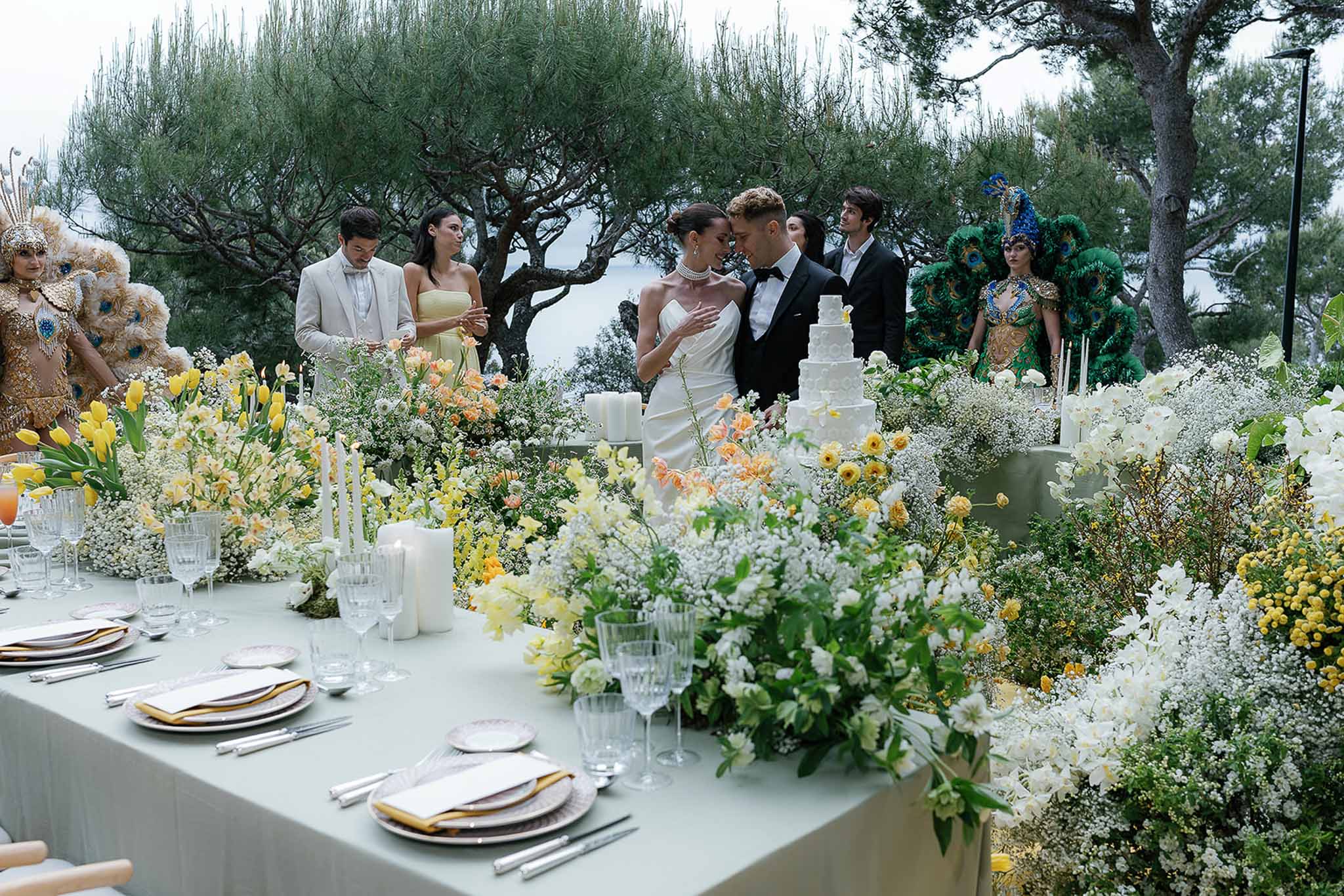 Bride and groom share intimate moment at outdoor reception table with yellow and white floral installation and tiered cake