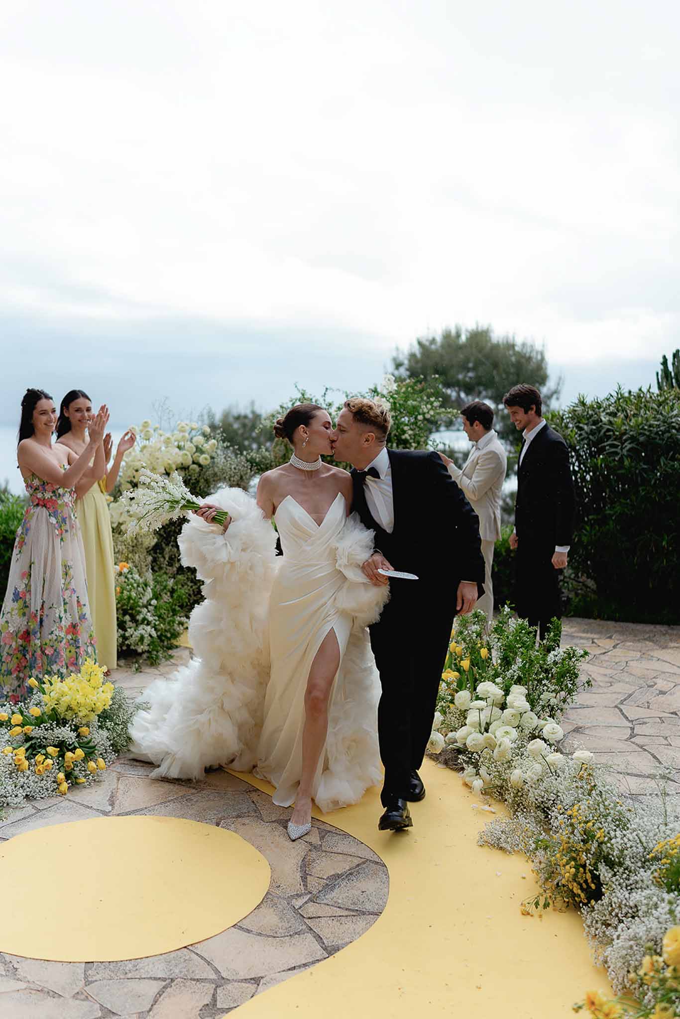 Bride and groom kiss walking up yellow aisle runner after outdoor seaside ceremony with wedding party