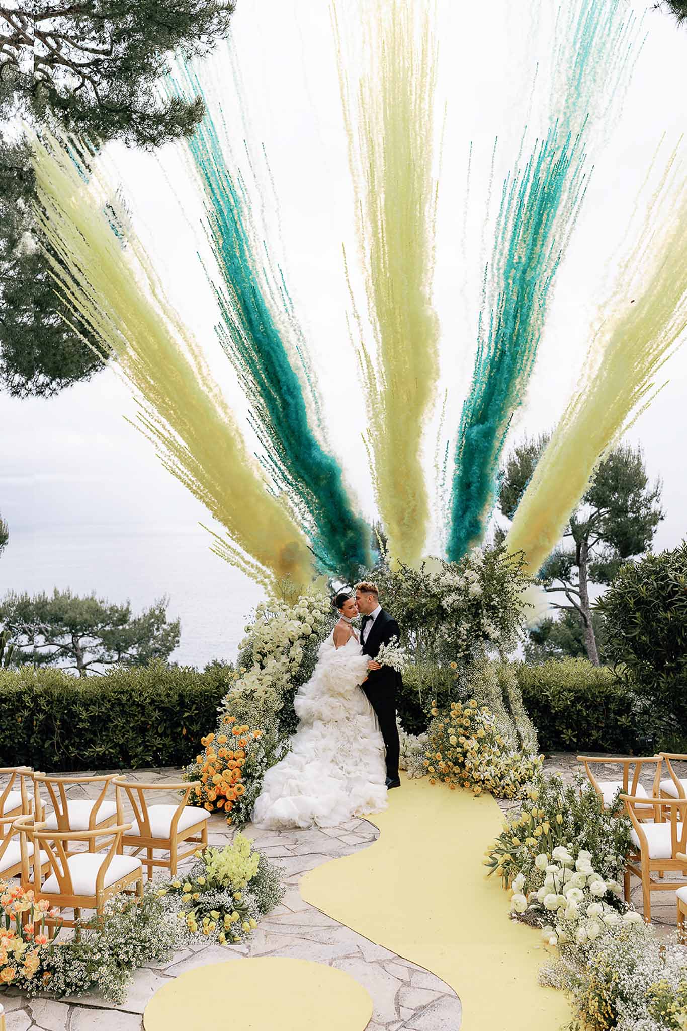 Couple kisses at altar under floral arch of hydrangeas and pampas grass with colored smoke cannons on coastal terrace