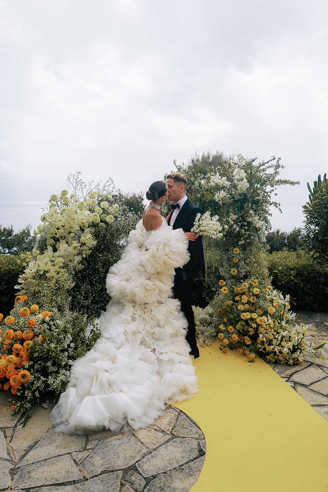 Bride and groom kiss at altar under floral arch with yellow aisle runner and sea view behind