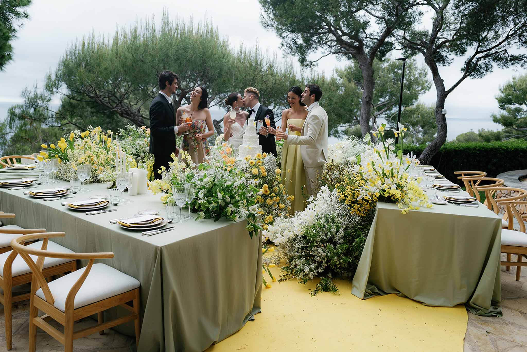 Outdoor terrace reception with sage green linens and yellow floral runners, couple kissing by wedding cake