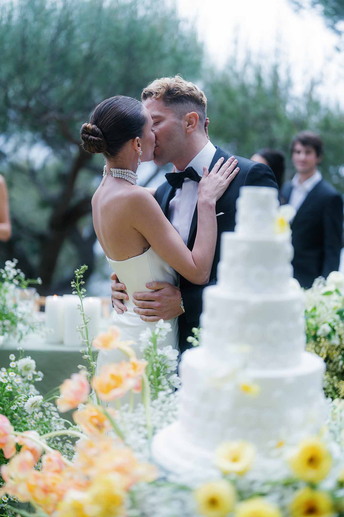 Bride and groom kiss behind five-tier white wedding cake surrounded by peach poppies and yellow ranunculus