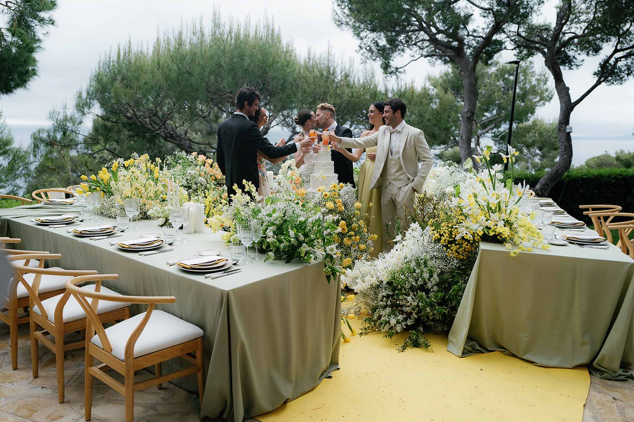 Guests toasting at sage-green table with yellow tulip runner, tiered cake, and sea view terrace behind