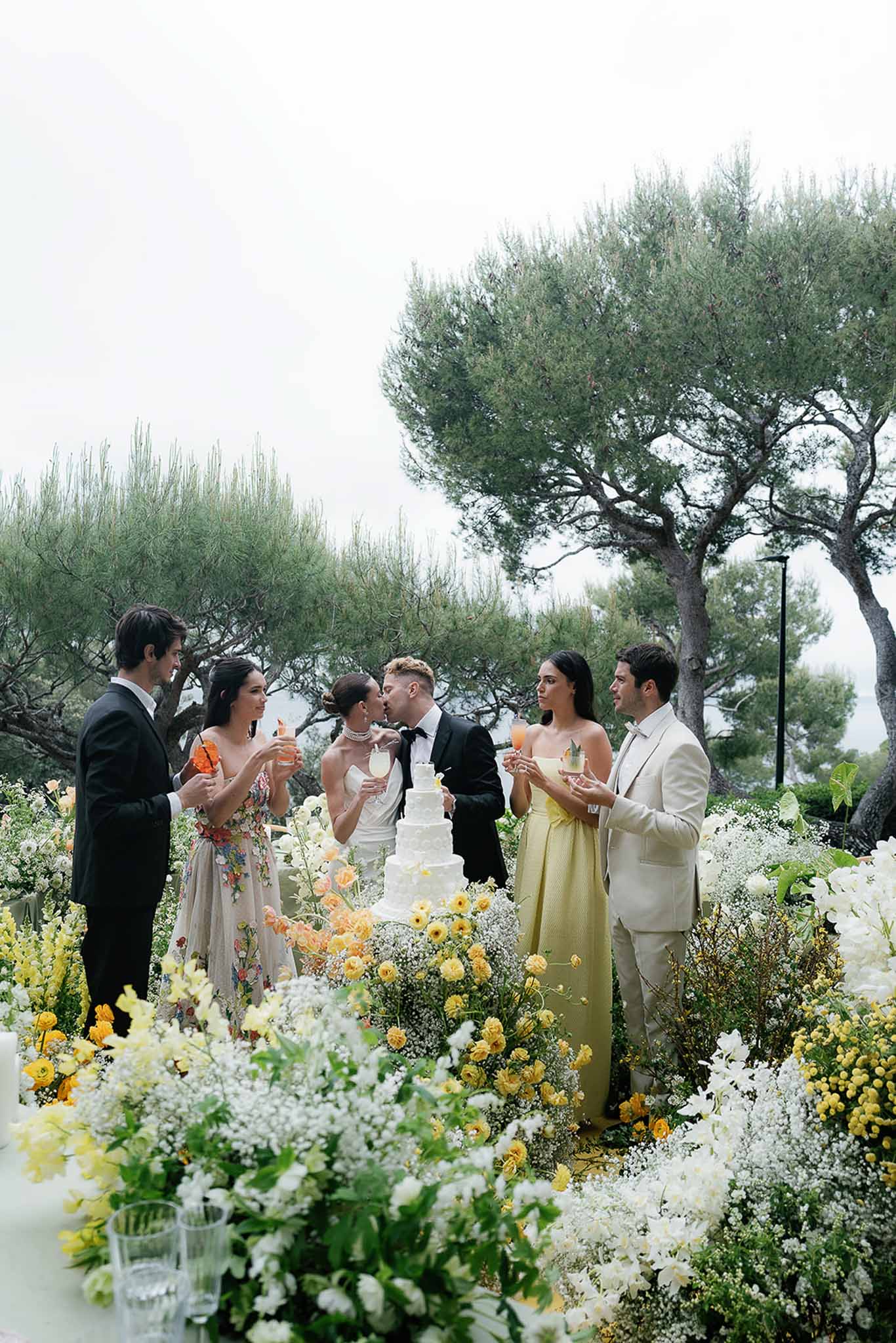 Couple kissing during cake cutting surrounded by guests and yellow dahlia floral installation