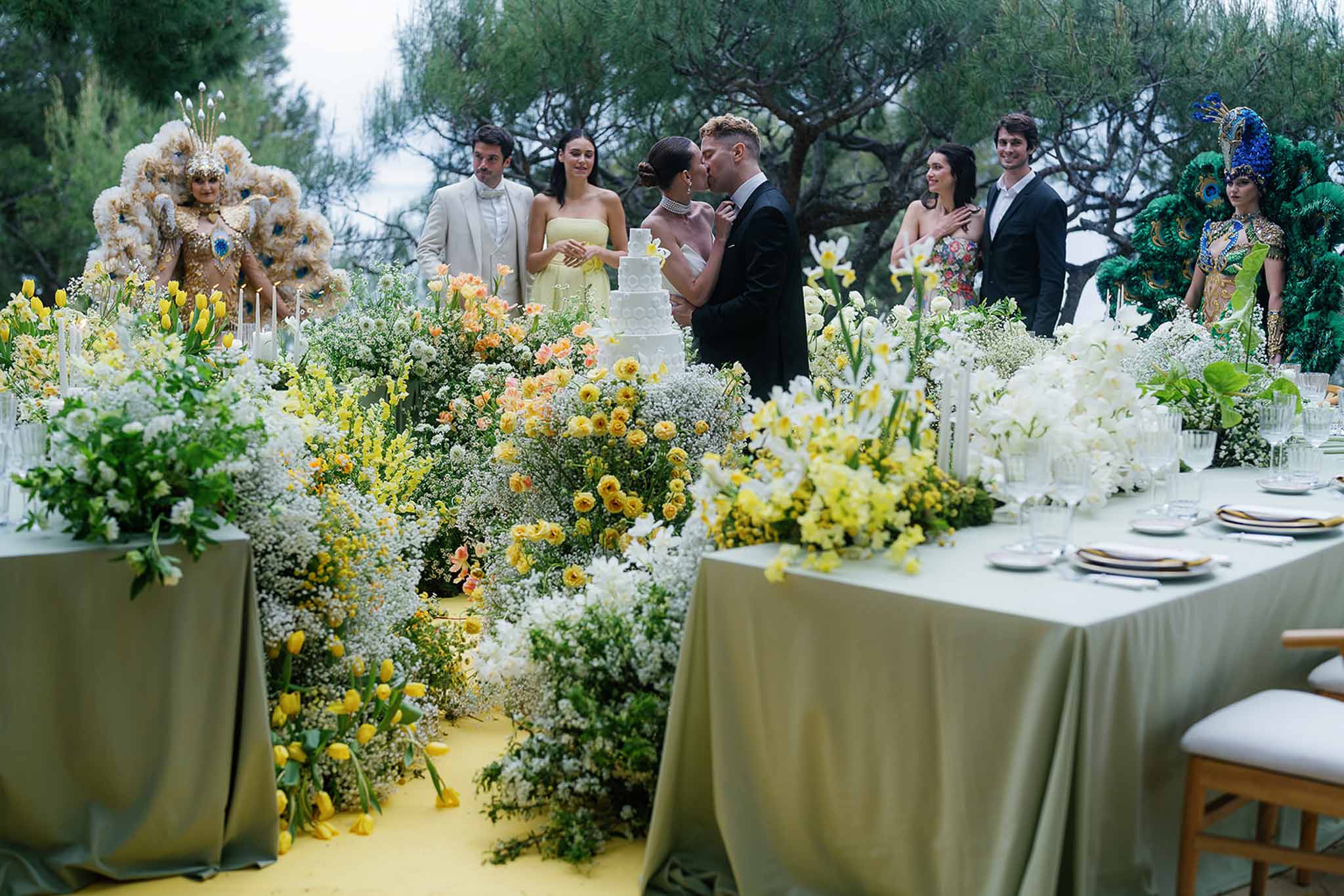 Bride and groom kiss during cake cutting surrounded by theatrical floral display and costumed performers outdoors