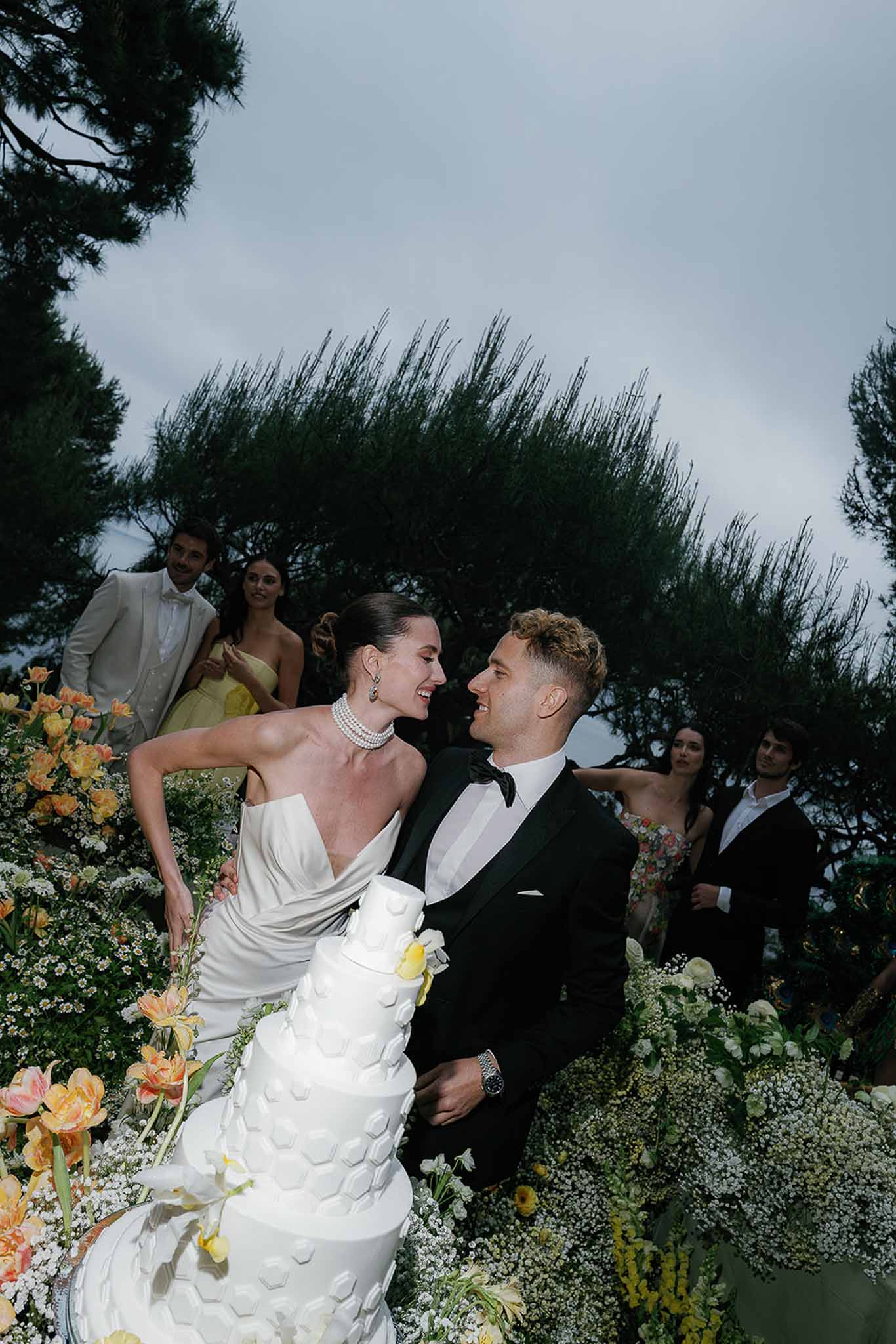 Bride and groom laughing while cutting four-tier honeycomb wedding cake surrounded by peach tulips and yellow blooms