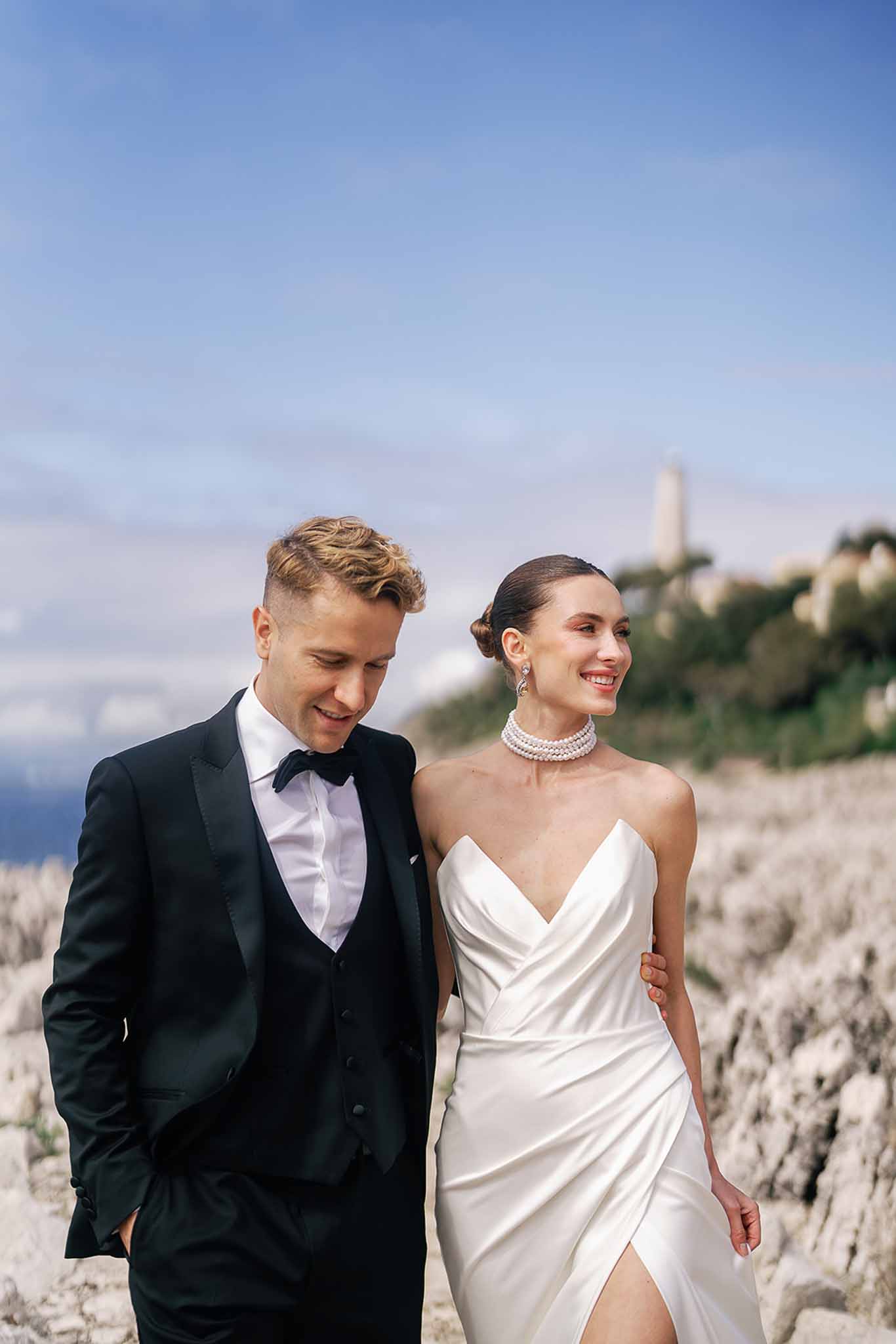 Bride in white satin slip gown with pearl choker and groom in black tuxedo smiling on rocky coastal clifftop