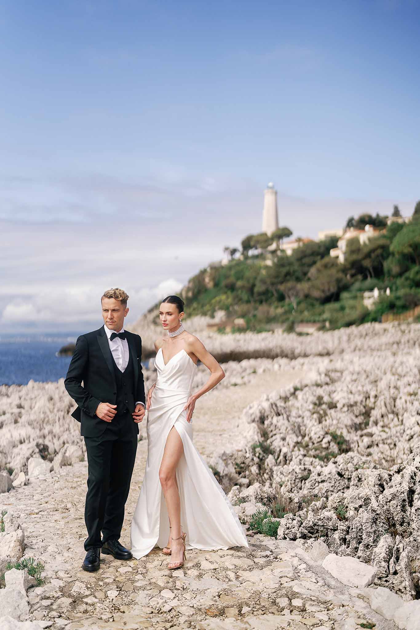 Couple on rocky coastal path with lighthouse behind, bride in ivory satin slip gown with pearl choker and gold heels