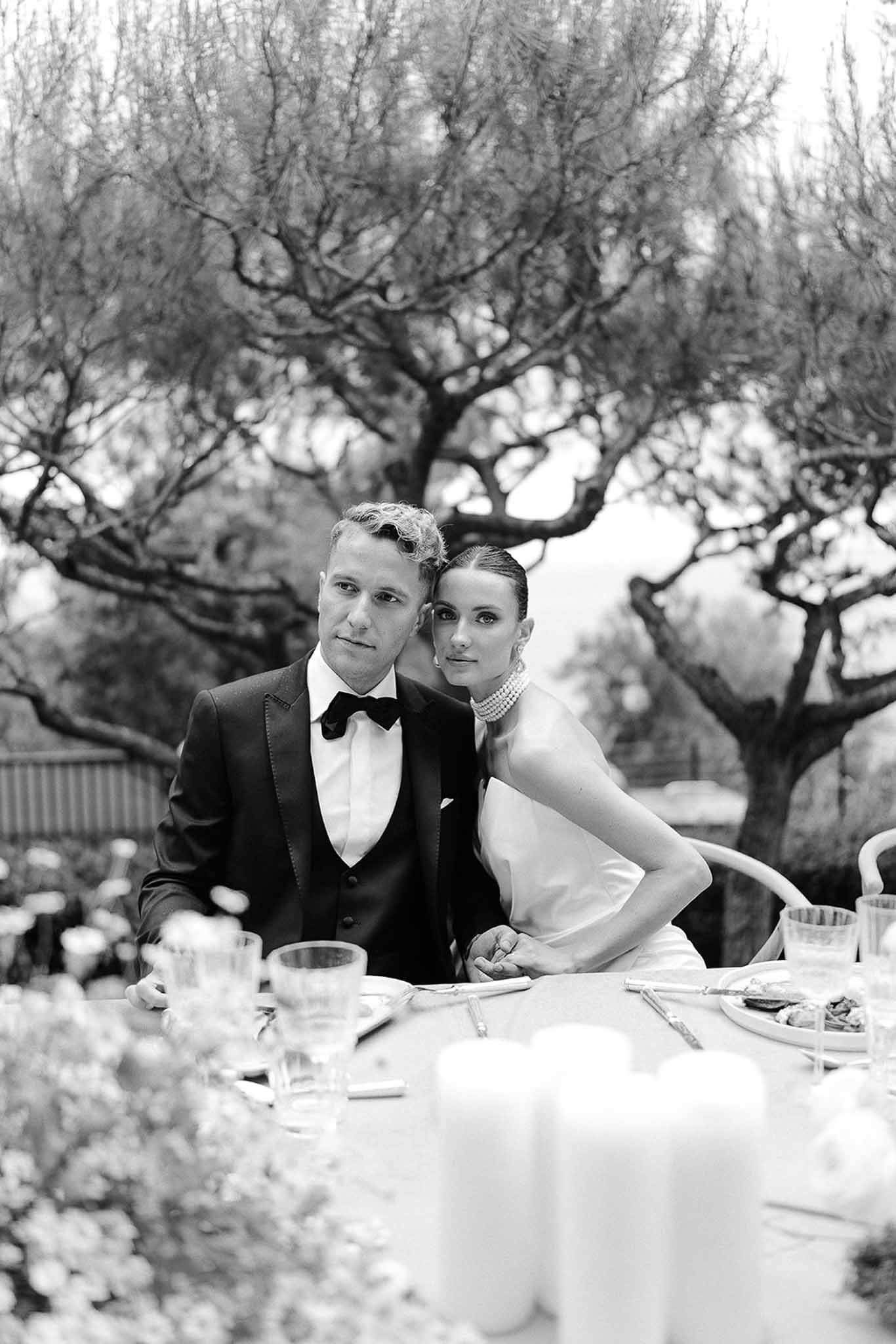 Black and white editorial portrait of couple at candlelit reception table, bride in pearl choker necklace