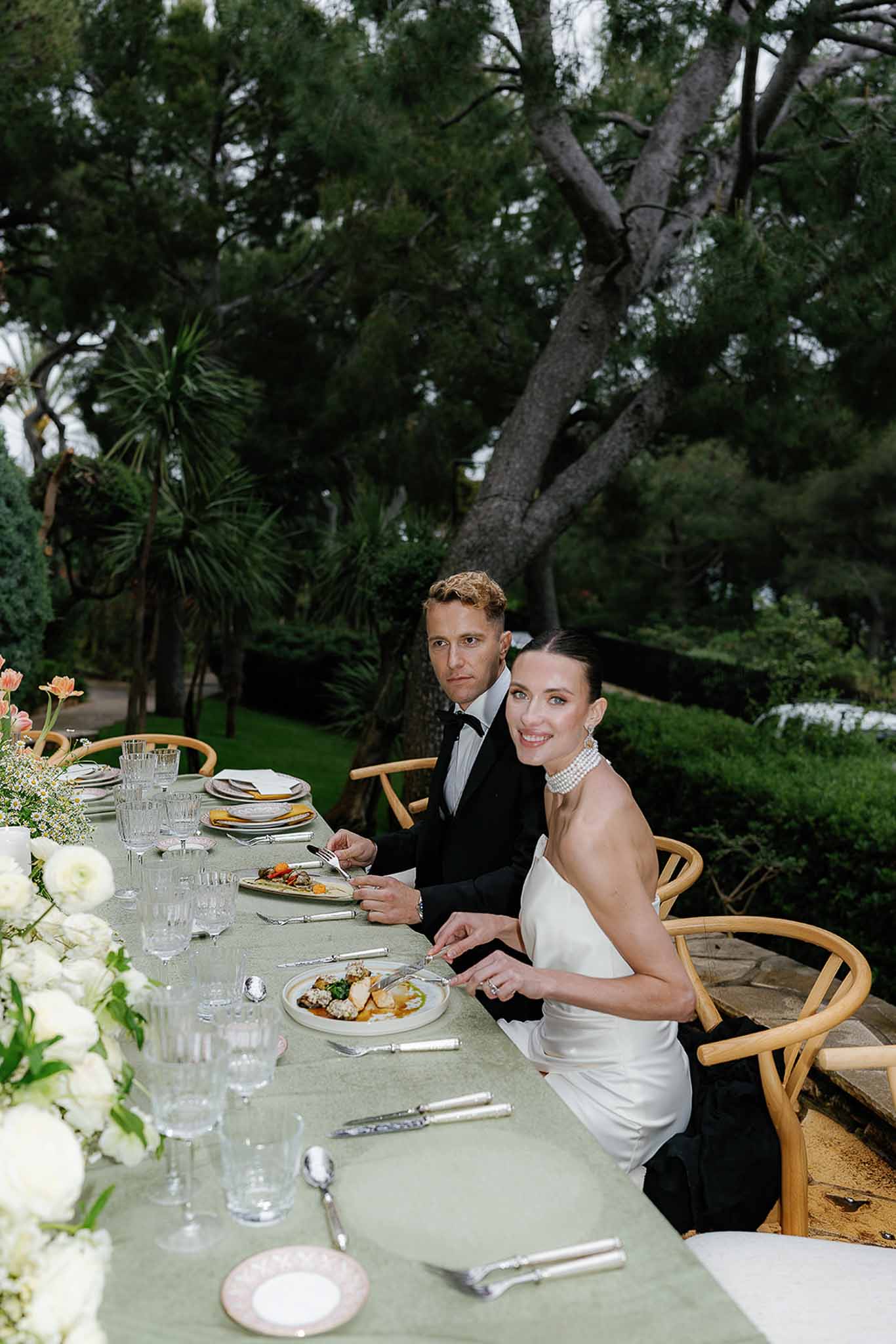 Bride and groom seated at outdoor reception table with sage linen and white floral centerpieces