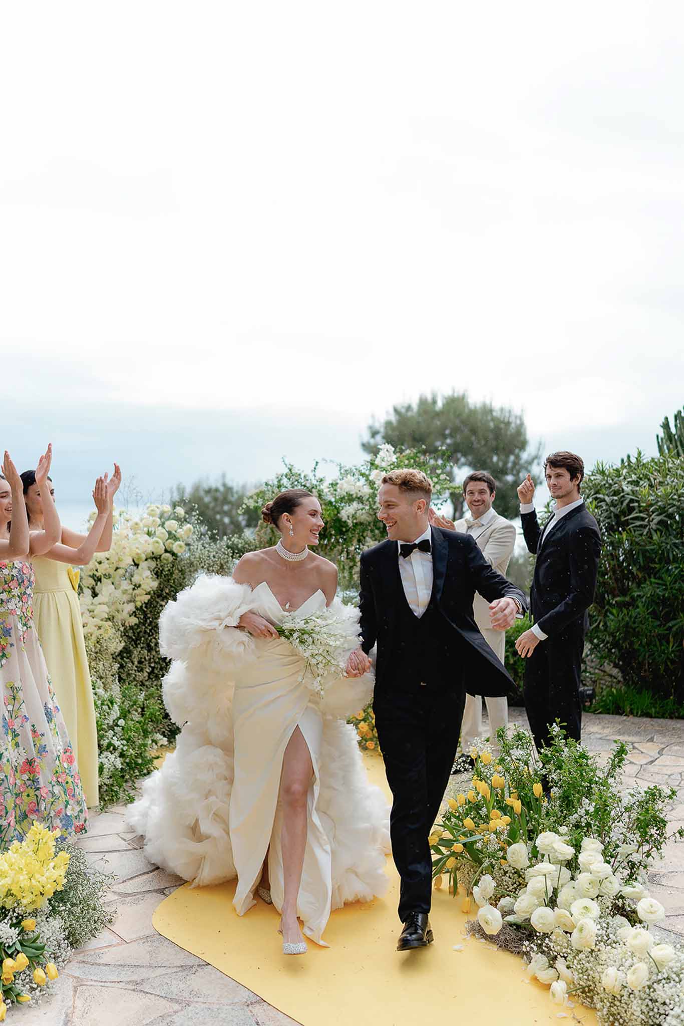 Bride with tulle wrap and groom walk yellow-runner aisle past yellow tulip and gypsophila arrangements