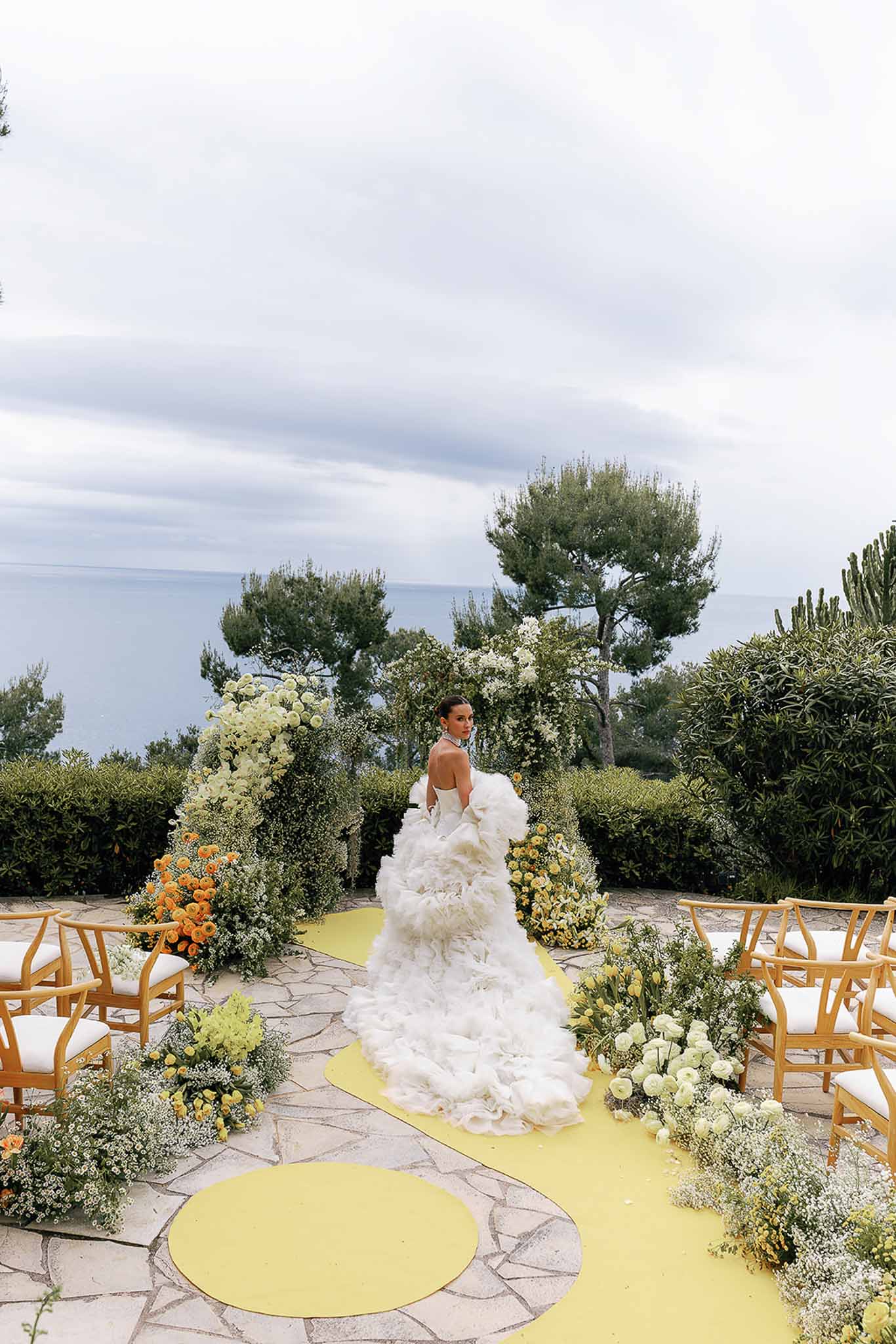 Bride at seaside altar with orchid arch, yellow aisle runner, and ranunculus arrangements on terrace