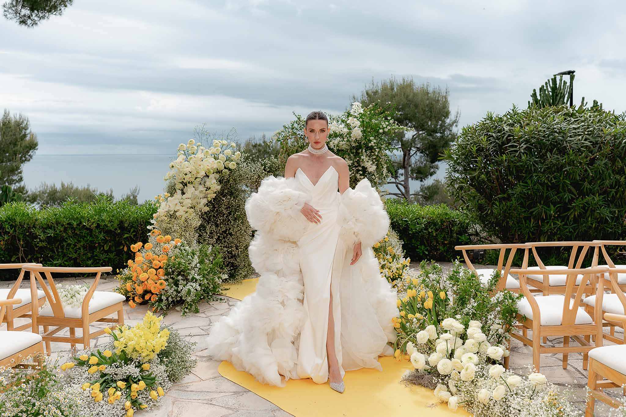 Bride in white satin gown with tulle cape standing at a yellow-and-white floral ceremony altar with sea view