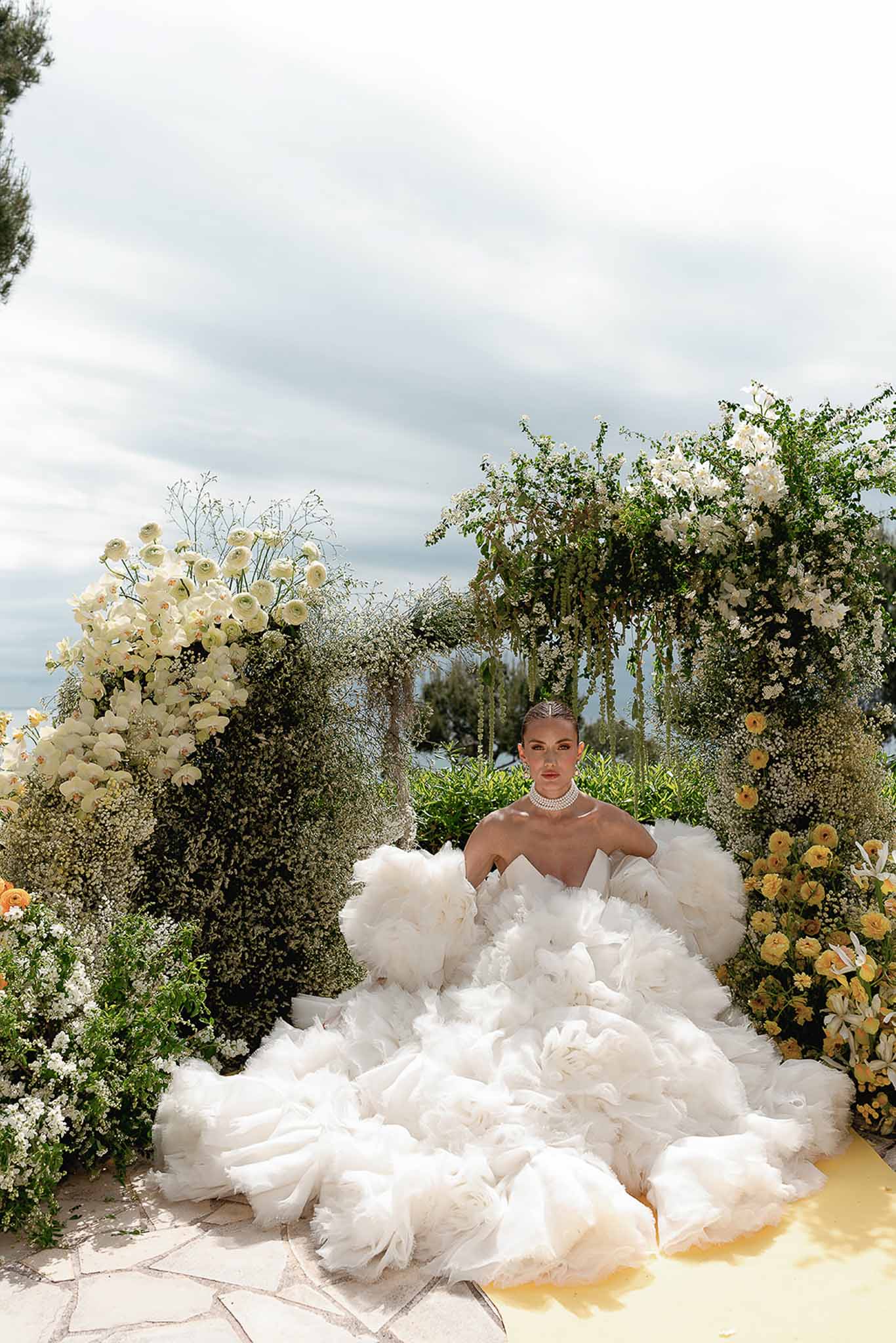 Bride in white tulle ballgown with pearl choker seated within white orchid and ranunculus floral arch