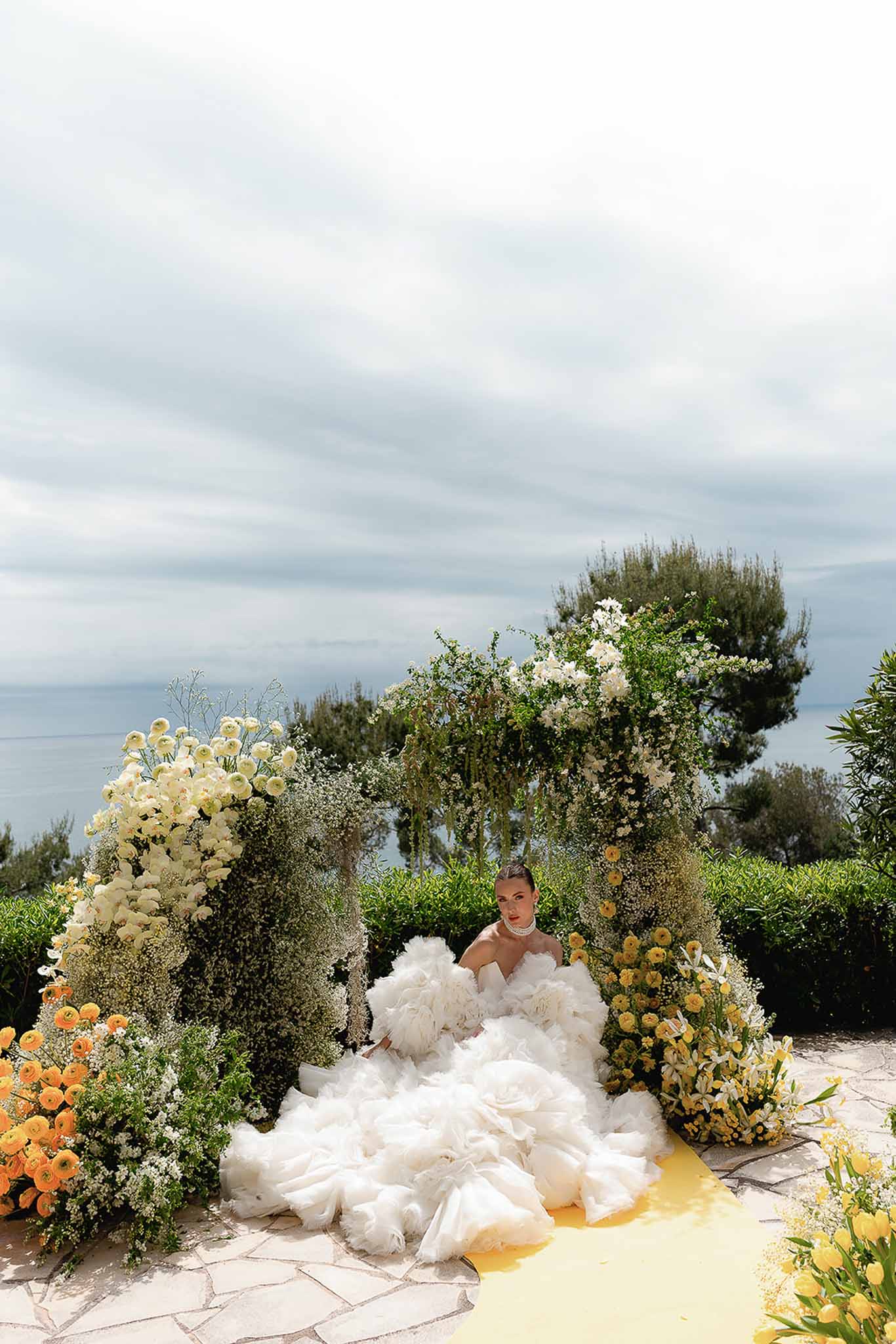Bride in ruffled ball gown with pearl choker amid orange yellow and white floral columns on sea-view terrace