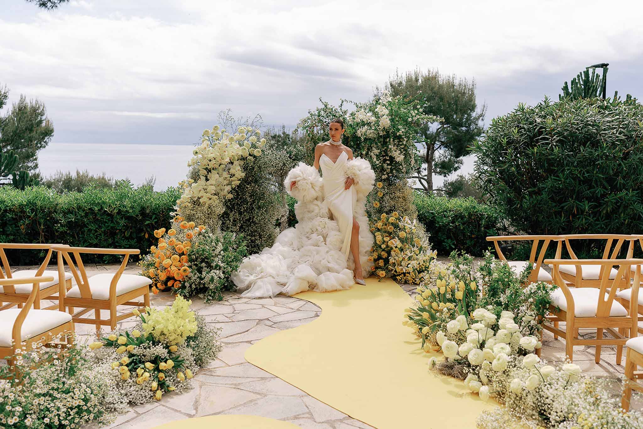 Bride at outdoor altar with yellow and orange floral aisle arrangements, greenery arch, and sea view backdrop