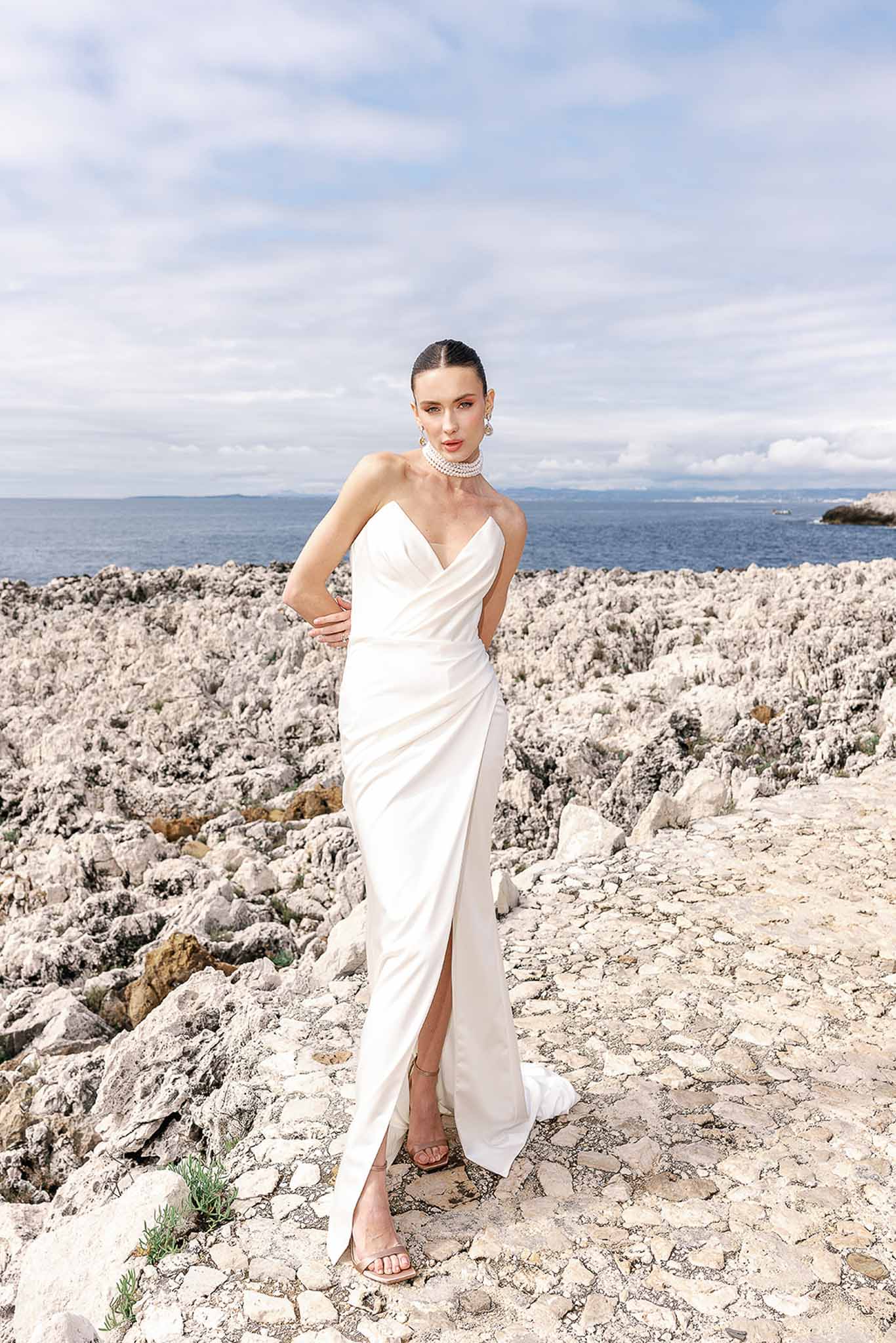 Bride in white strapless column gown with pearl choker posing on rocky Mediterranean shoreline