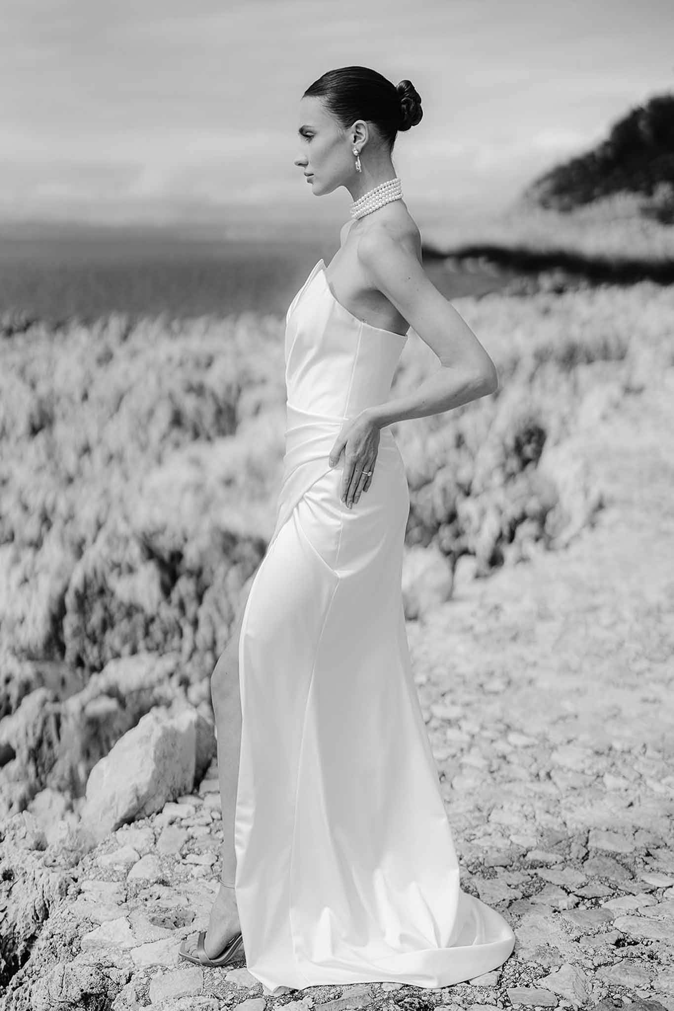 Black-and-white full-length bridal portrait of woman in strapless column gown with pearl choker on rocky coastline