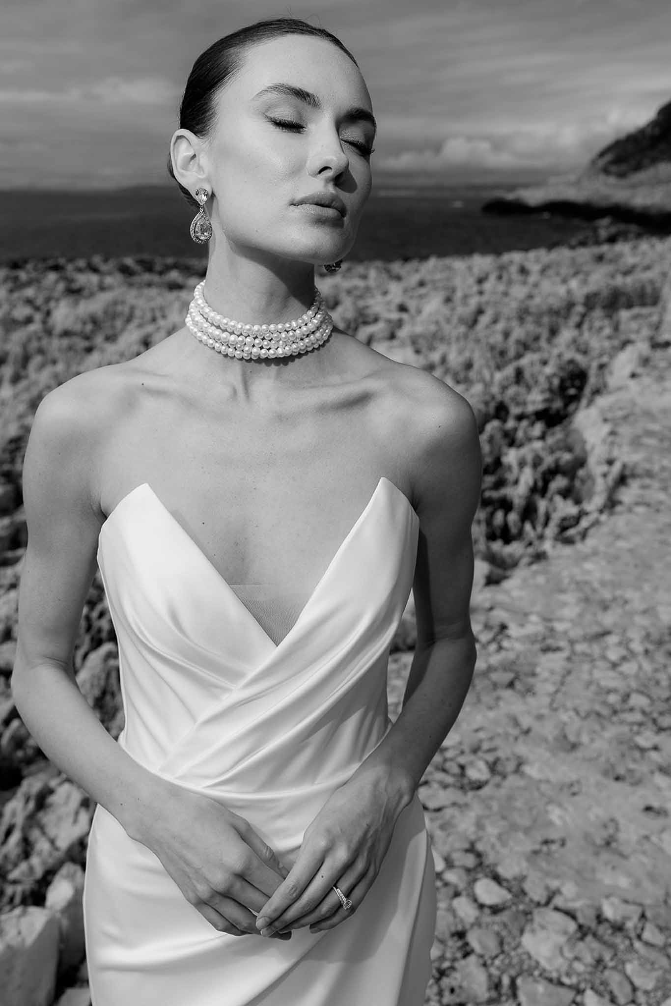 Black and white coastal portrait of bride in strapless satin gown with pearl choker and drop earrings