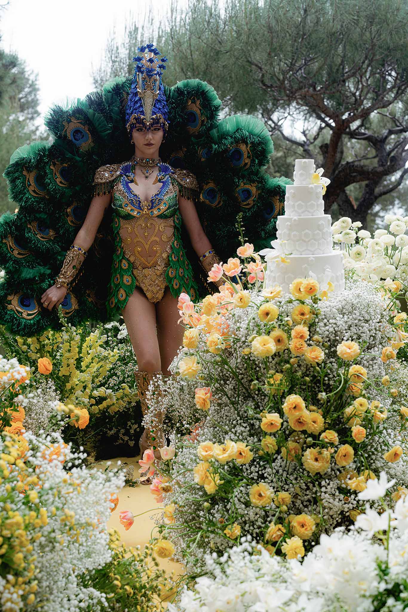 Peacock-costumed performer walking past five-tier wedding cake and yellow ranunculus floral installations