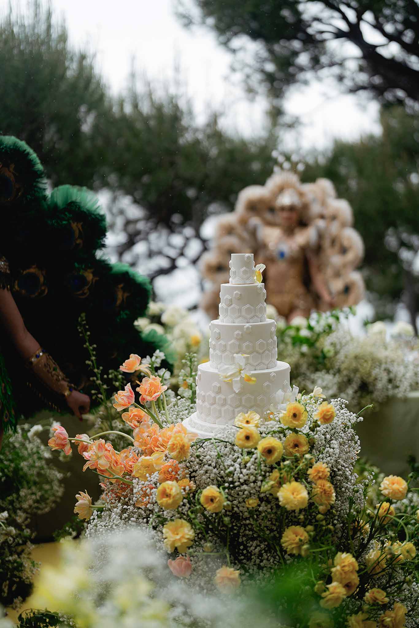 Five-tier hexagonal white cake surrounded by yellow ranunculus and coral parrot tulips with carnival performers