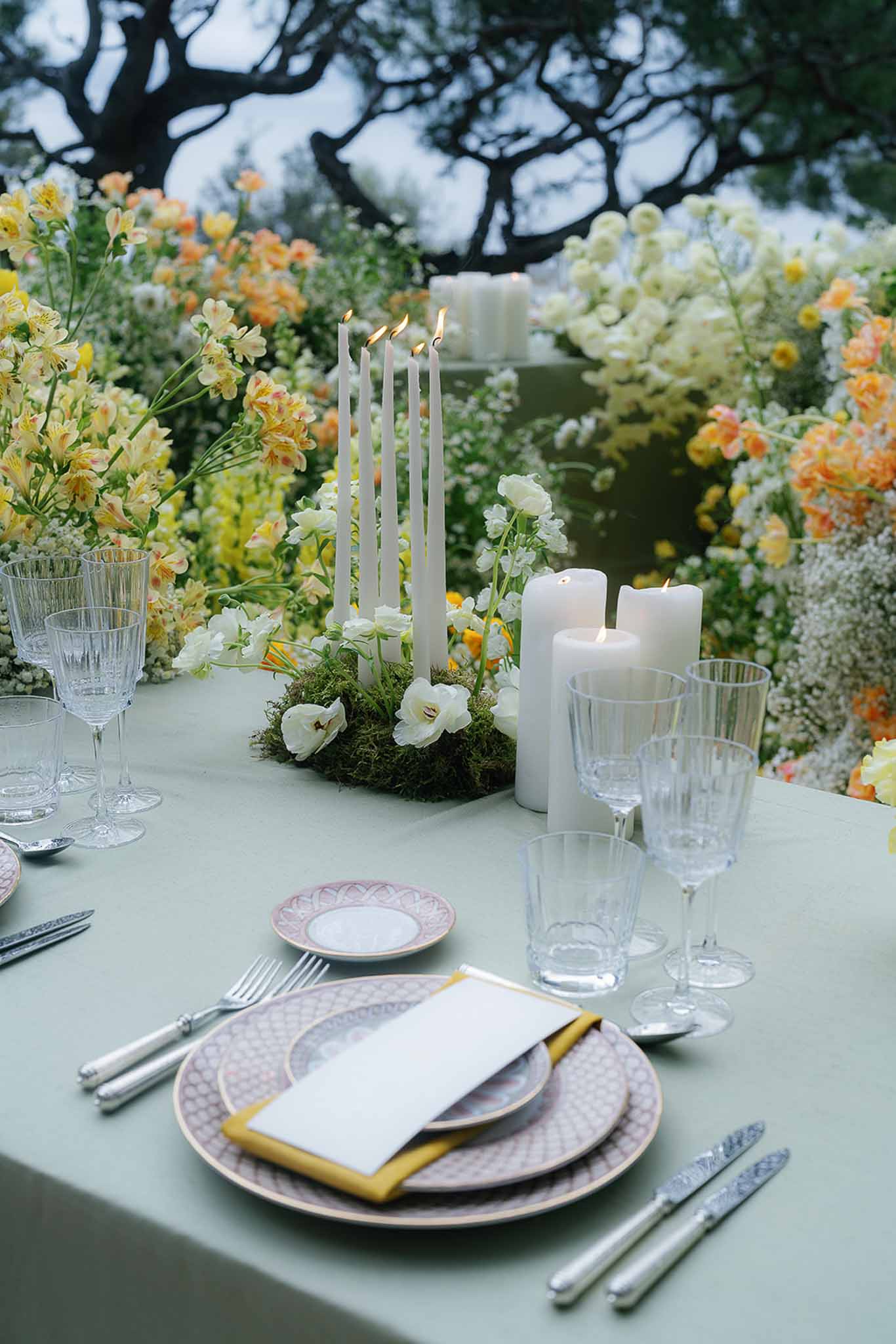 Place setting with patterned plates, mustard napkin, and sage linen beside yellow and peach floral backdrop with lit tapers