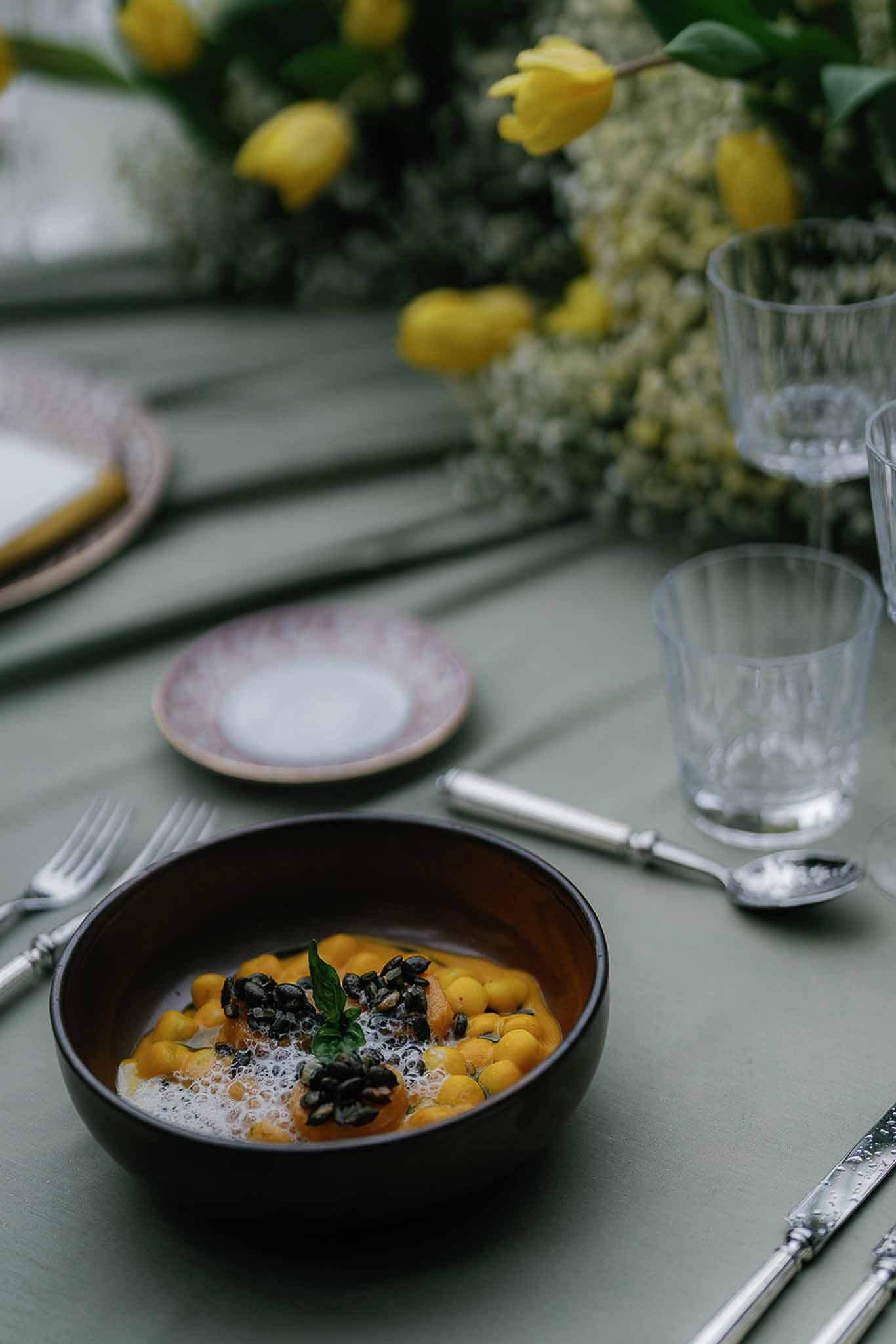 Wedding reception place setting with orange dish in dark bowl, sage green linen, crystal glassware, and yellow tulip centr...