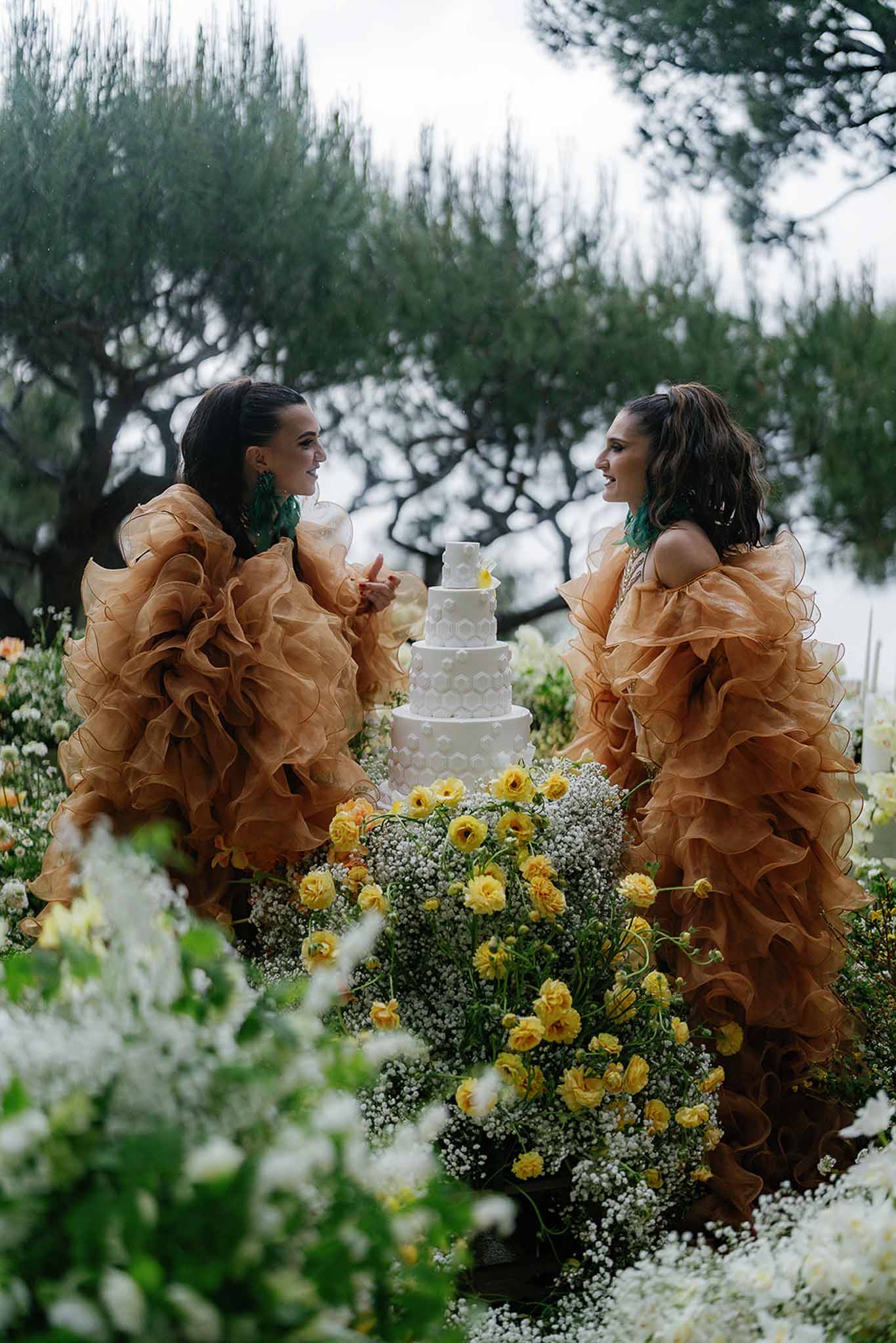 Two brides in amber ruffled gowns beside honeycomb-textured cake with yellow ranunculus ground flowers