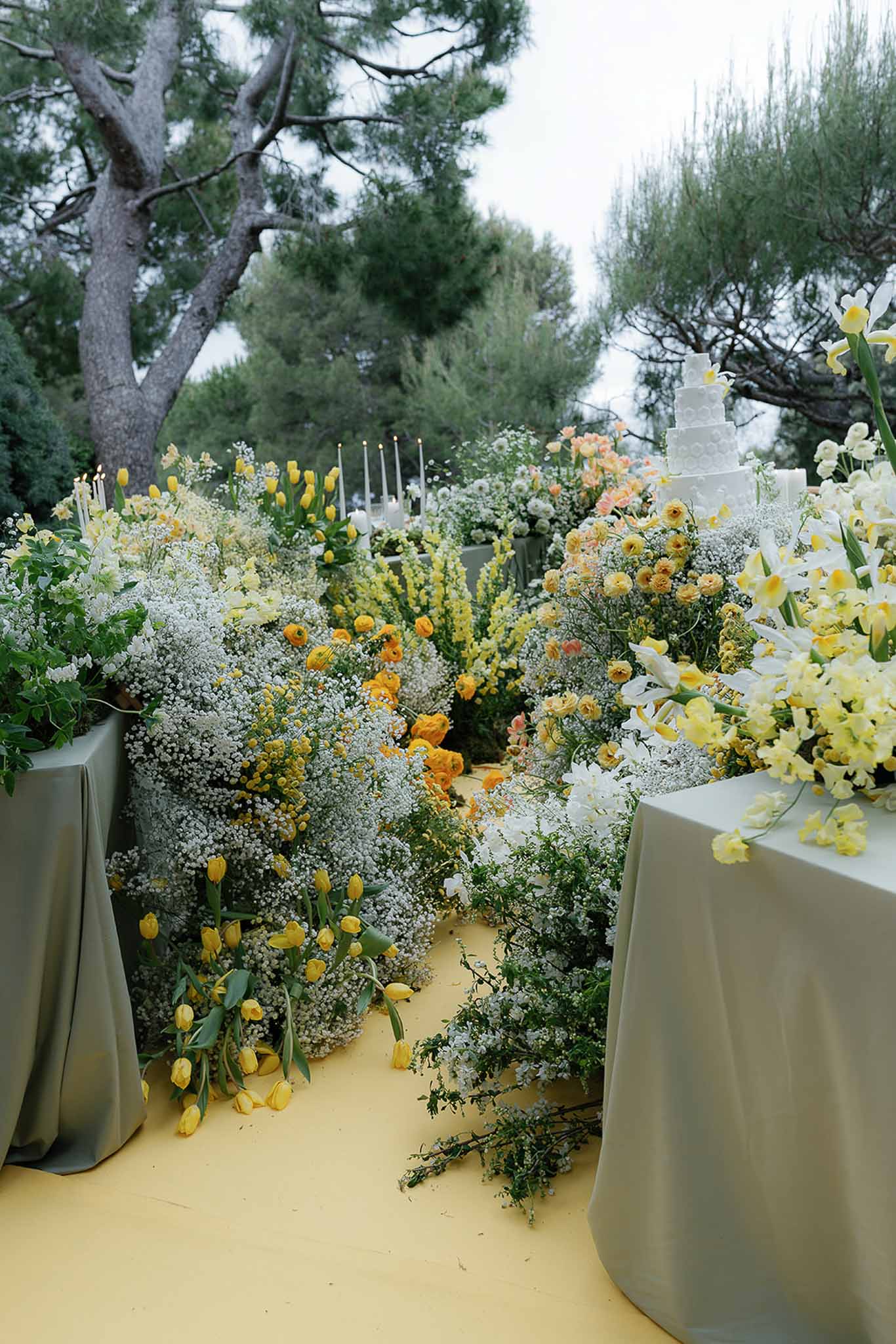 Garden-style floral installation with yellow tulips and ranunculus surrounding white tiered wedding cake on sage green linens