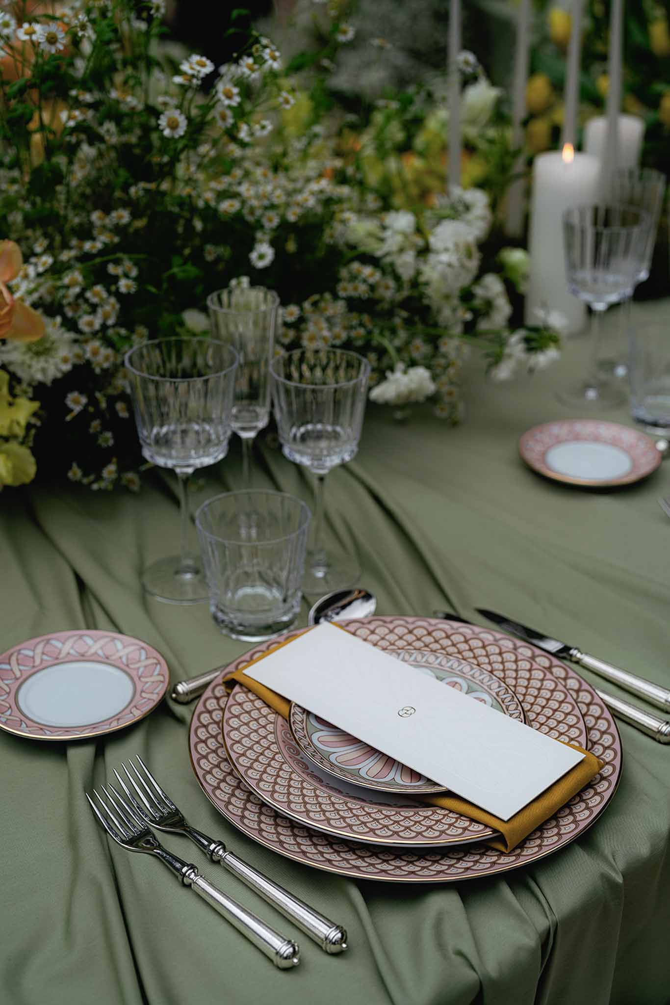 Reception place setting with patterned plates, yellow napkin, and menu on sage green linen with daisy centerpiece