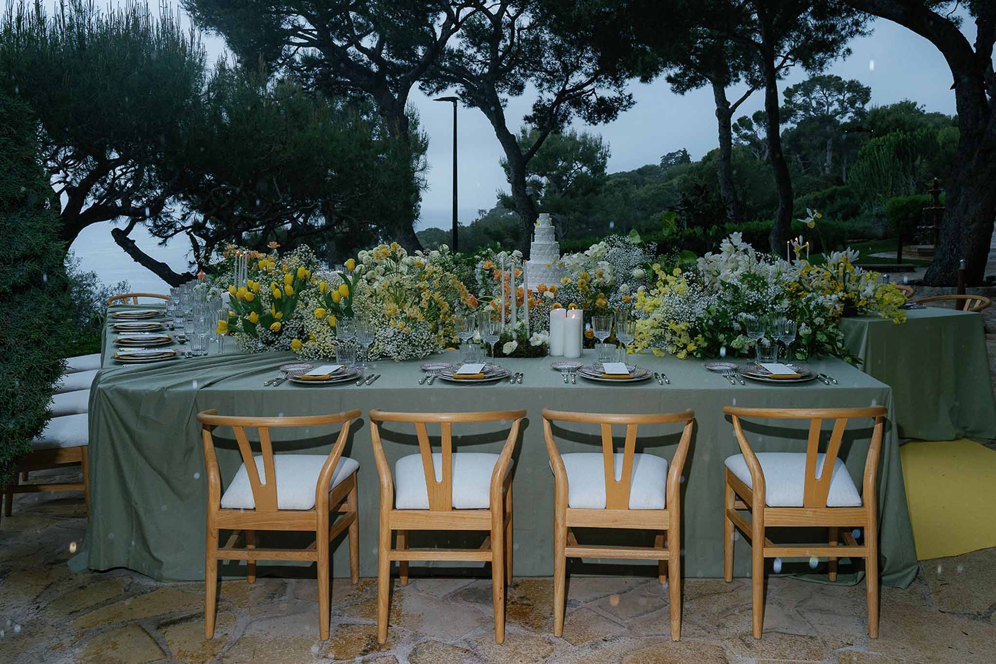 Dusk reception table with sage green linen, yellow tulips, white orchids, and tiered cake flanked by pillar candles on ter...