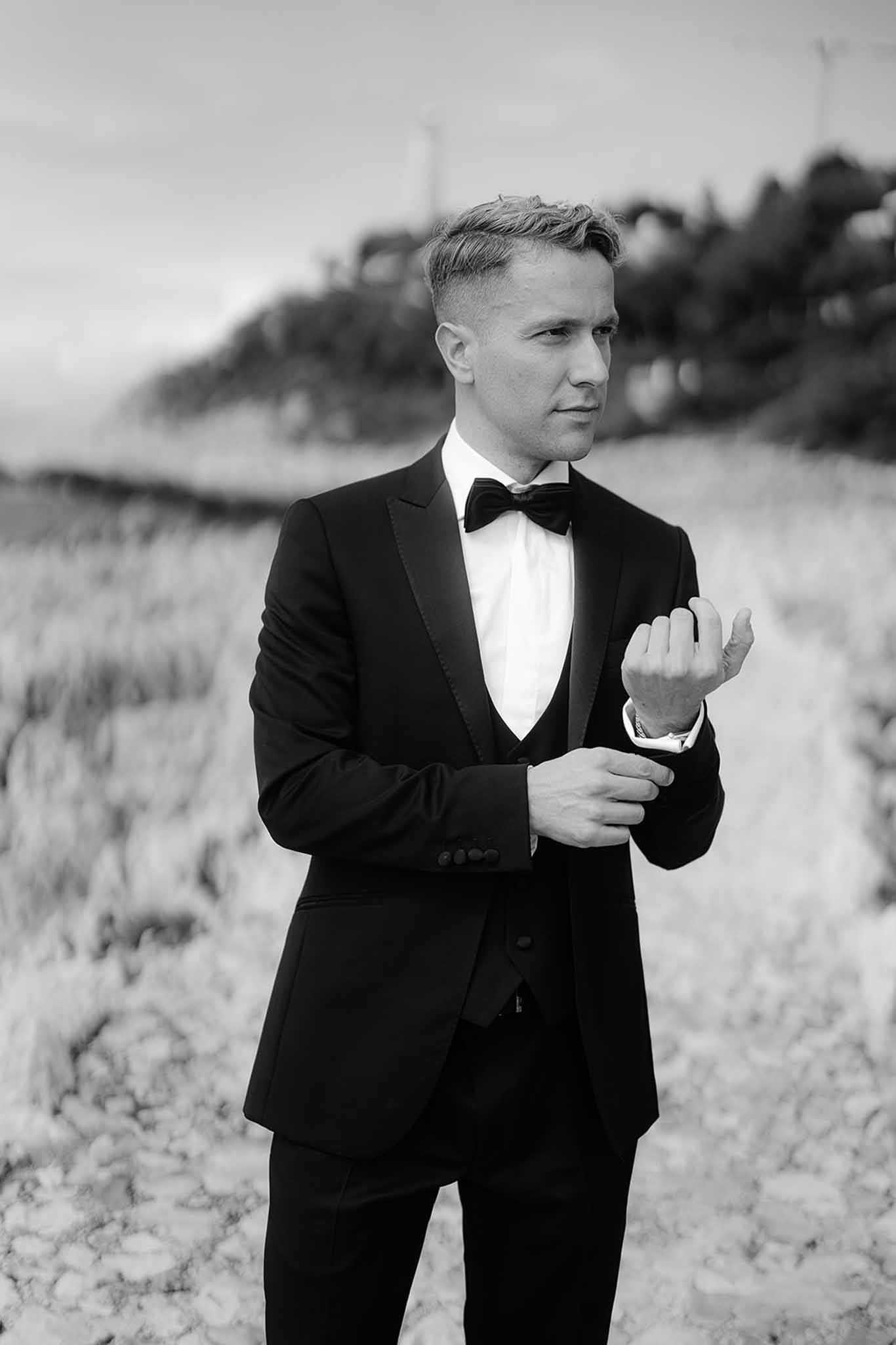Black and white groom adjusting cufflinks in tuxedo with satin lapels bow tie outdoors with hillside behind
