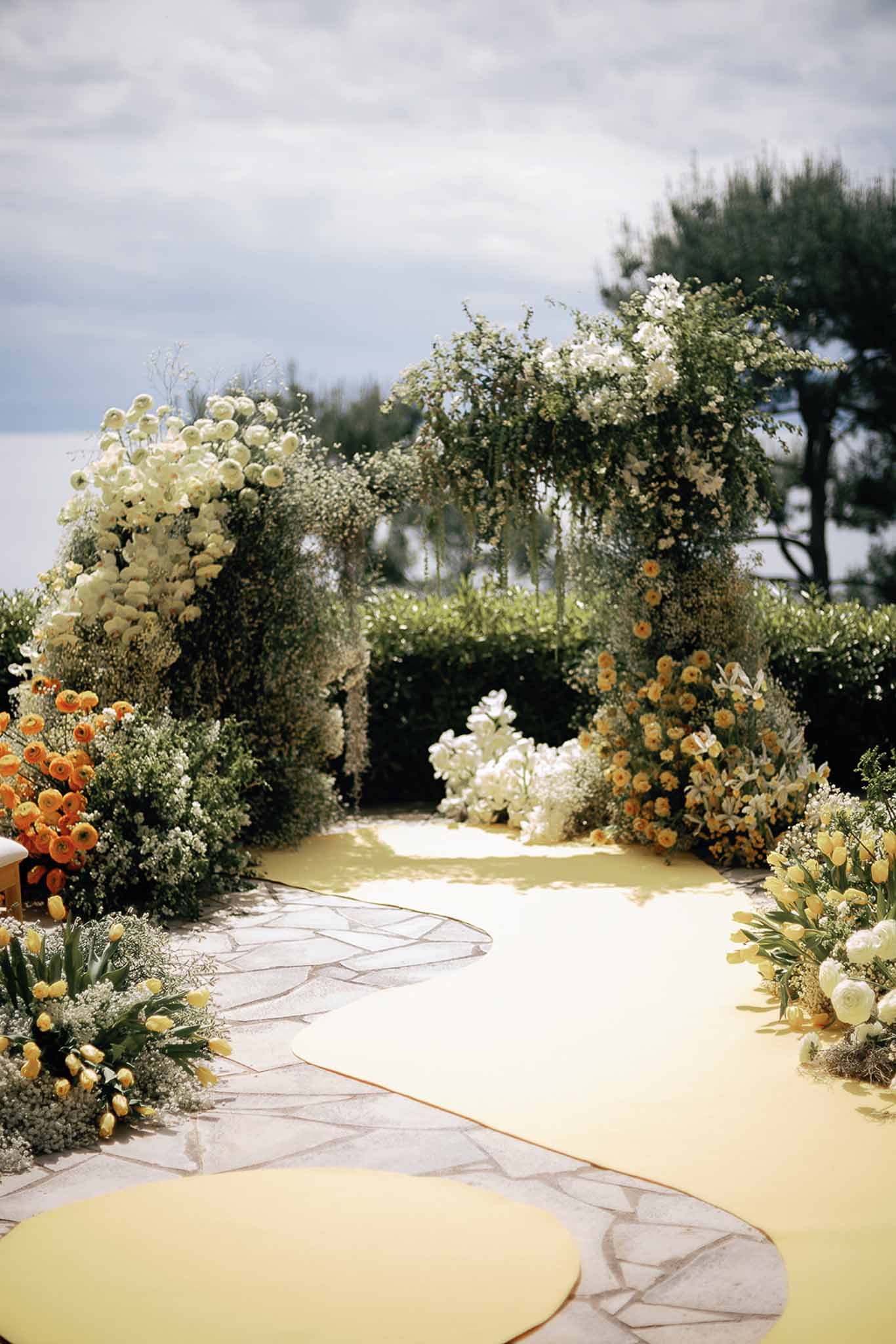 Outdoor ceremony setup with greenery floral arch, yellow aisle runner, and orange and white ranunculus arrangements on sto...