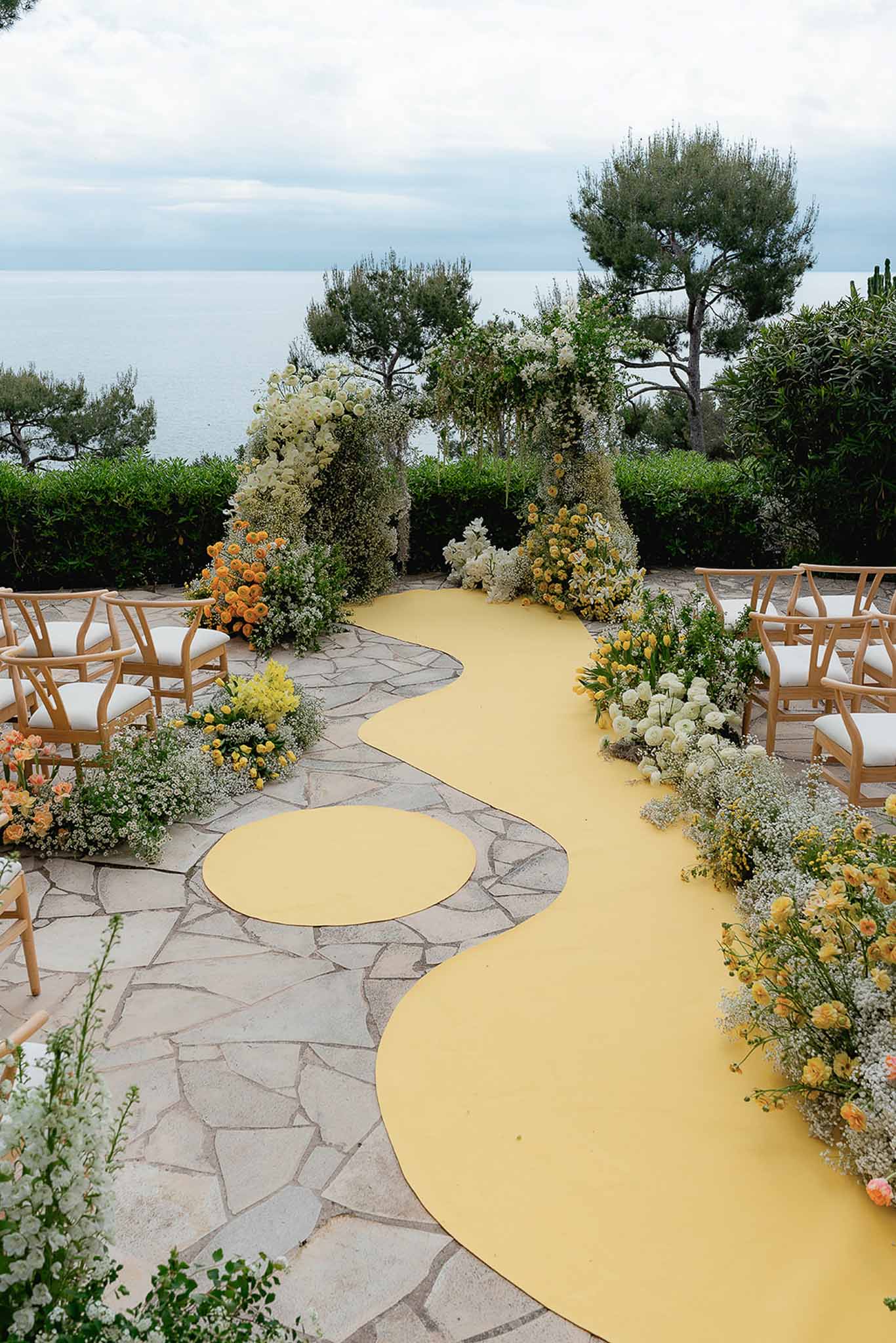 Seaside ceremony with yellow sculptural aisle runner, floral arch, and wishbone chairs overlooking the sea