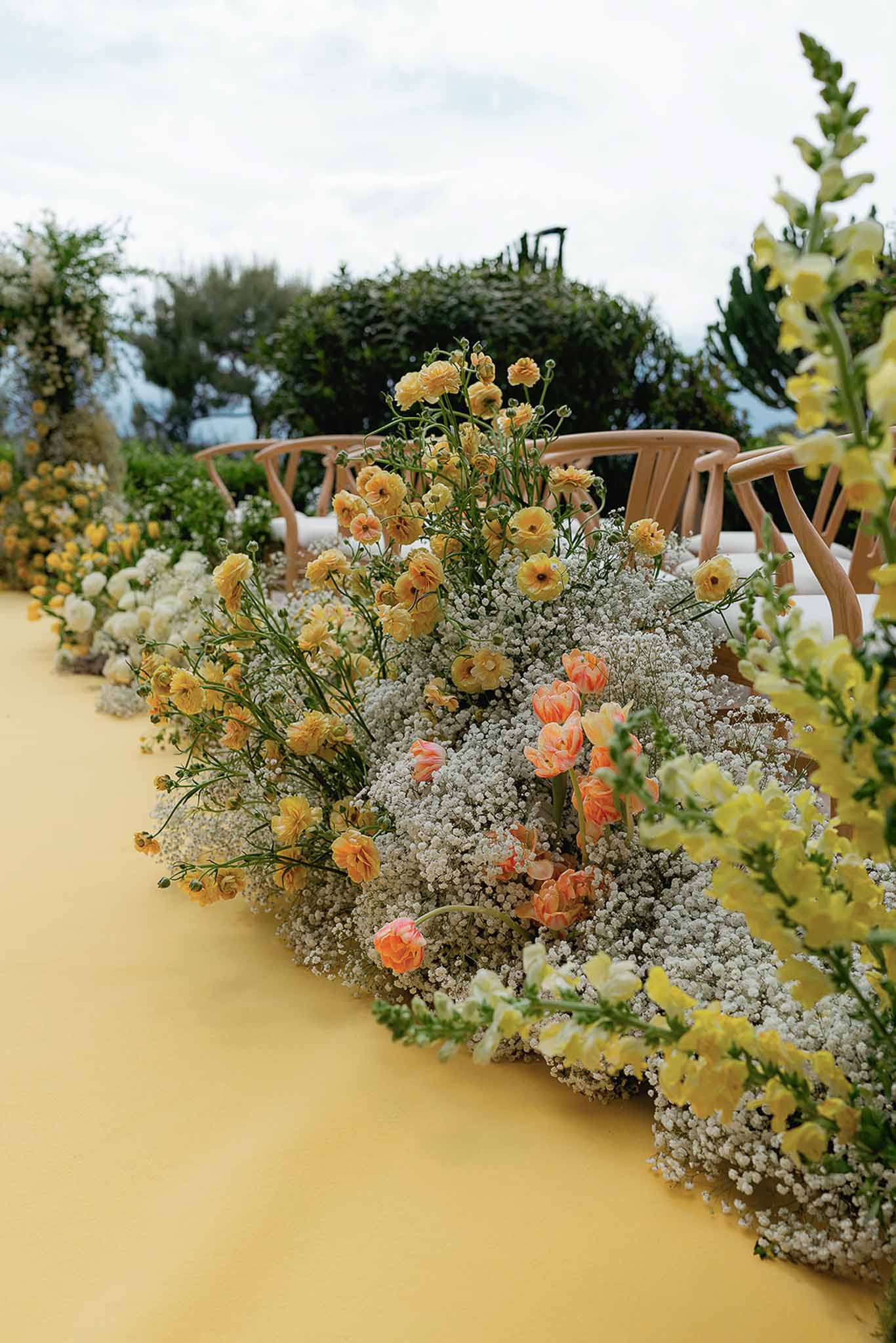 Yellow ceremony aisle runner bordered by wild garden-style flowers with ranunculus, tulips, and baby's breath