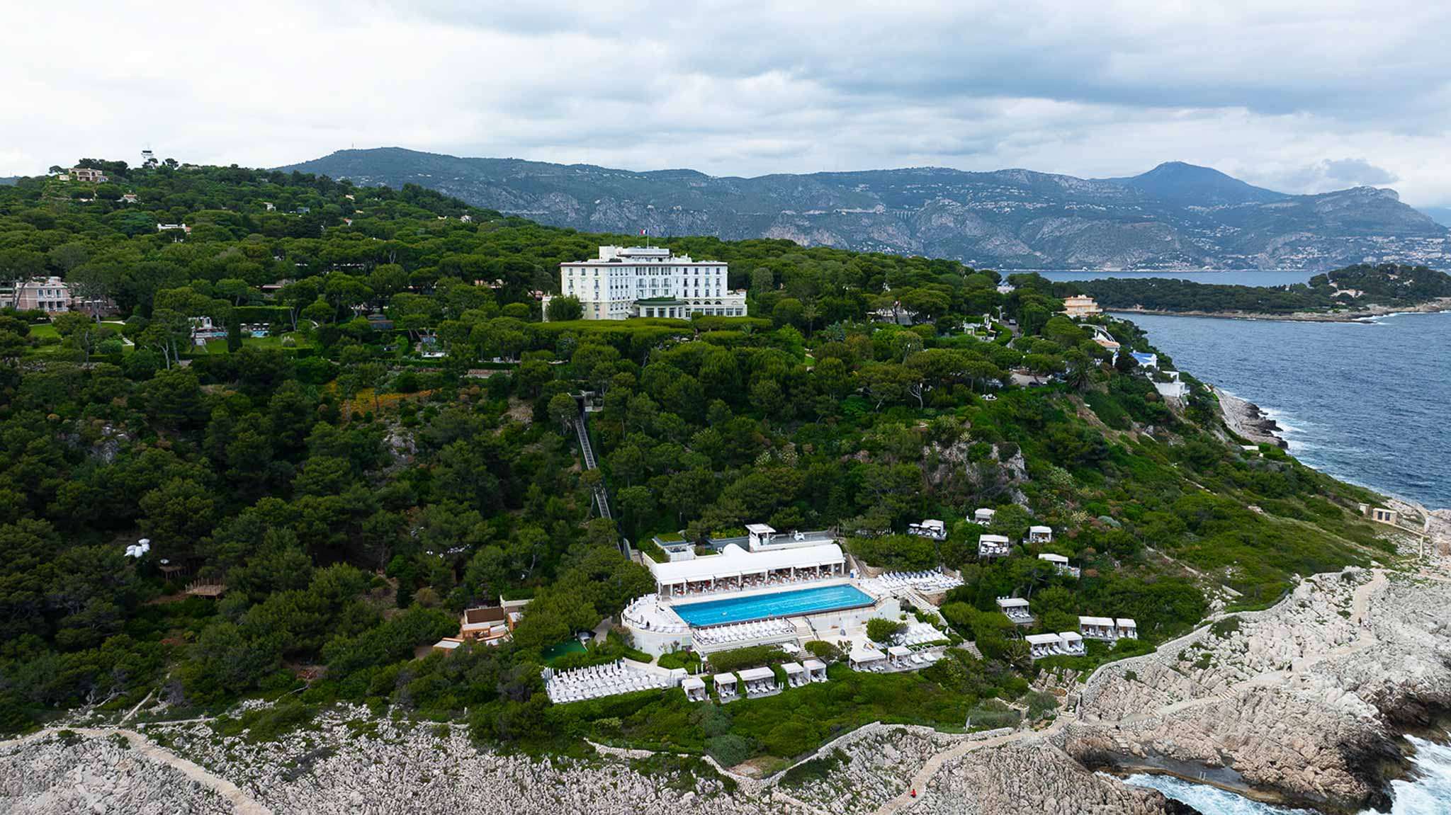Aerial view of white Belle Epoque hotel on rocky French Riviera headland with turquoise pool and coastal cliffs