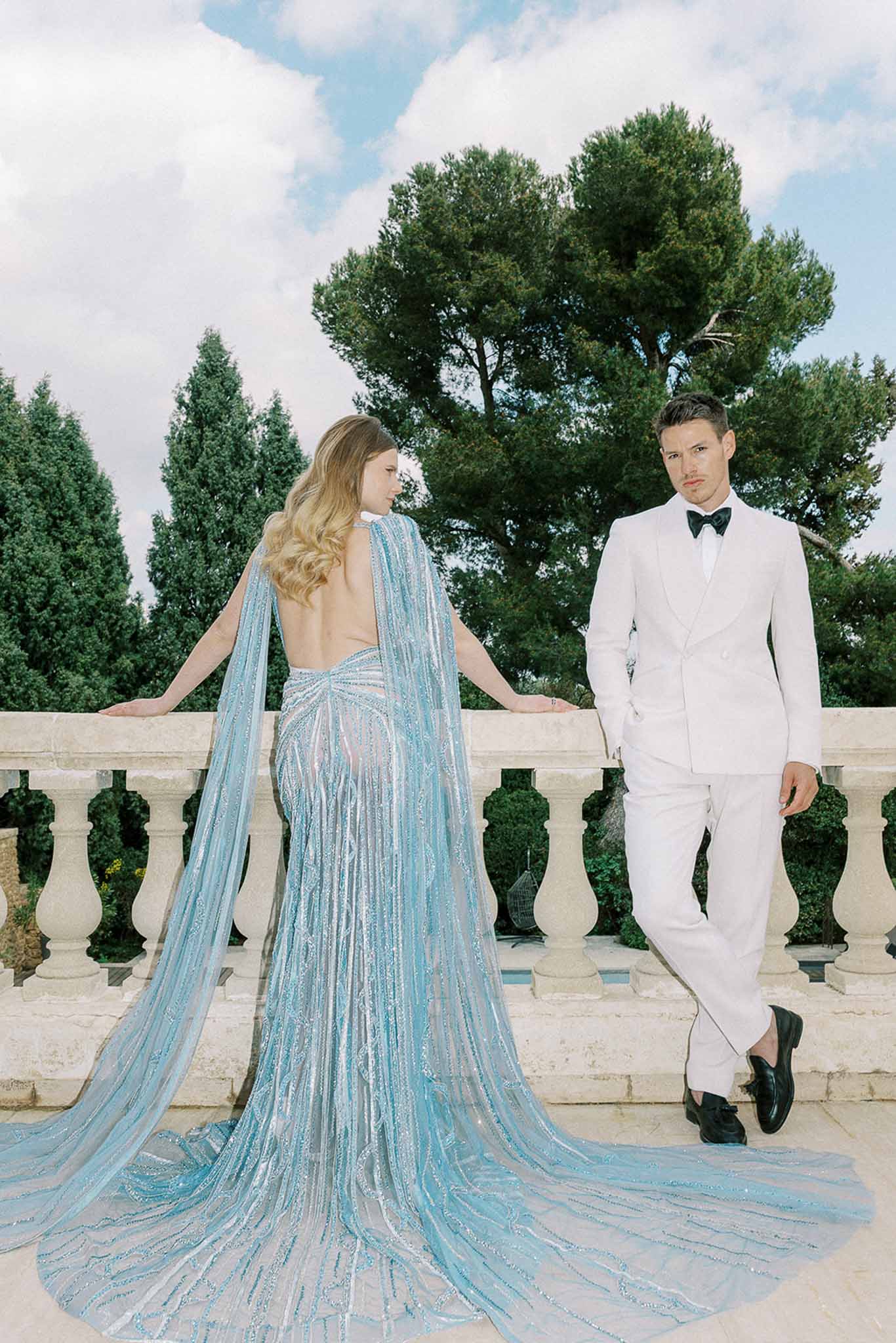 Couple on stone balustrade terrace with bride in blue sequined cape gown and groom in white suit