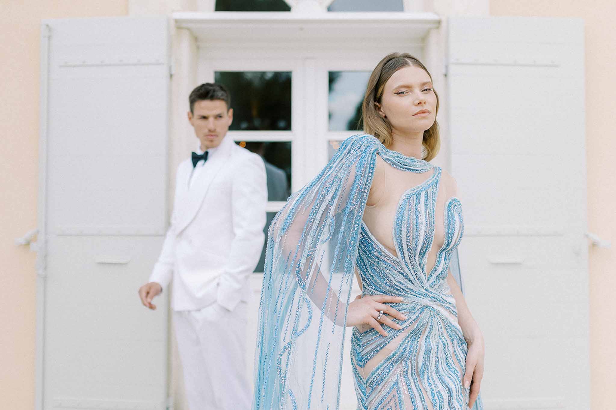 Bride in sky blue beaded gown with sheer cape and groom in white tuxedo before peach building